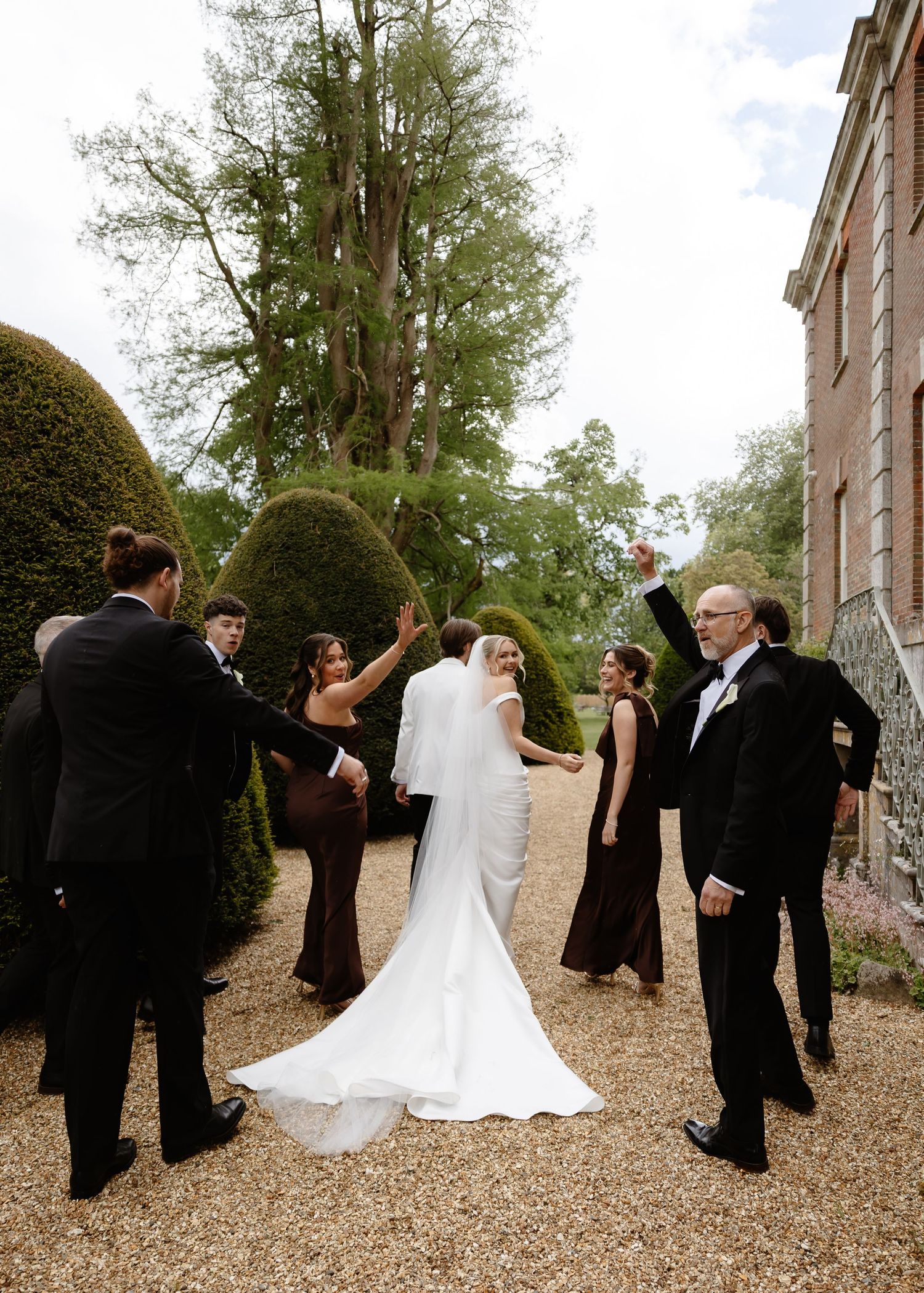 Bride in white gown with veil celebrates with wedding party in dark attire amid manicured hedges at elegant estate.