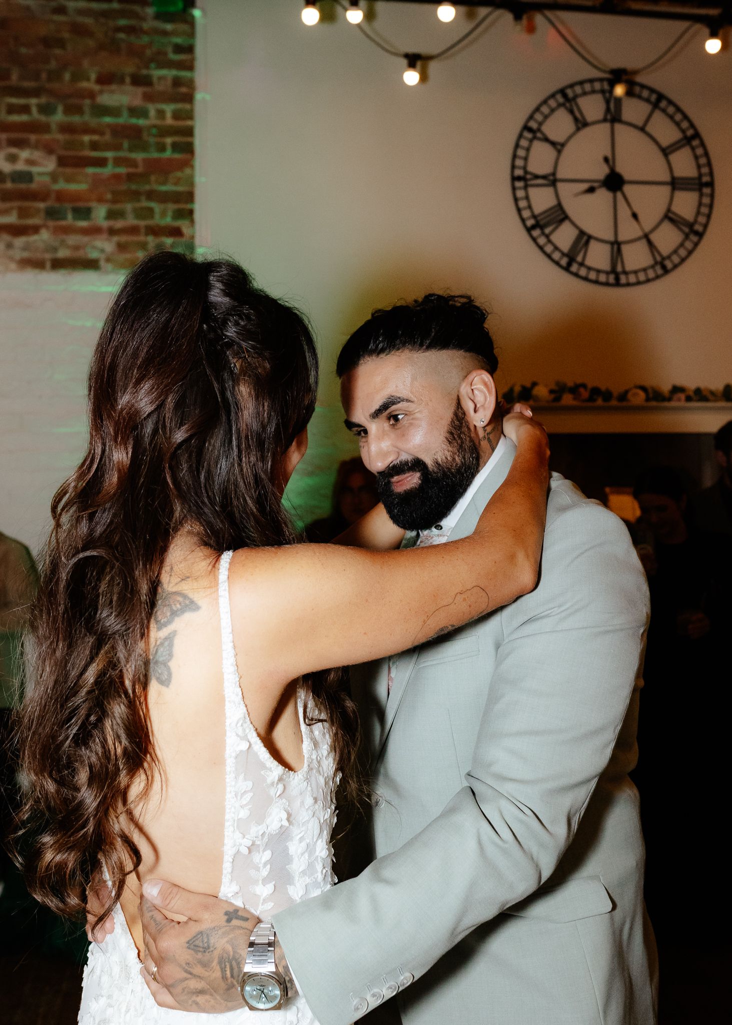 A couple in wedding attire shares a romantic dance moment under string lights in a rustic brick-walled venue with a clock.