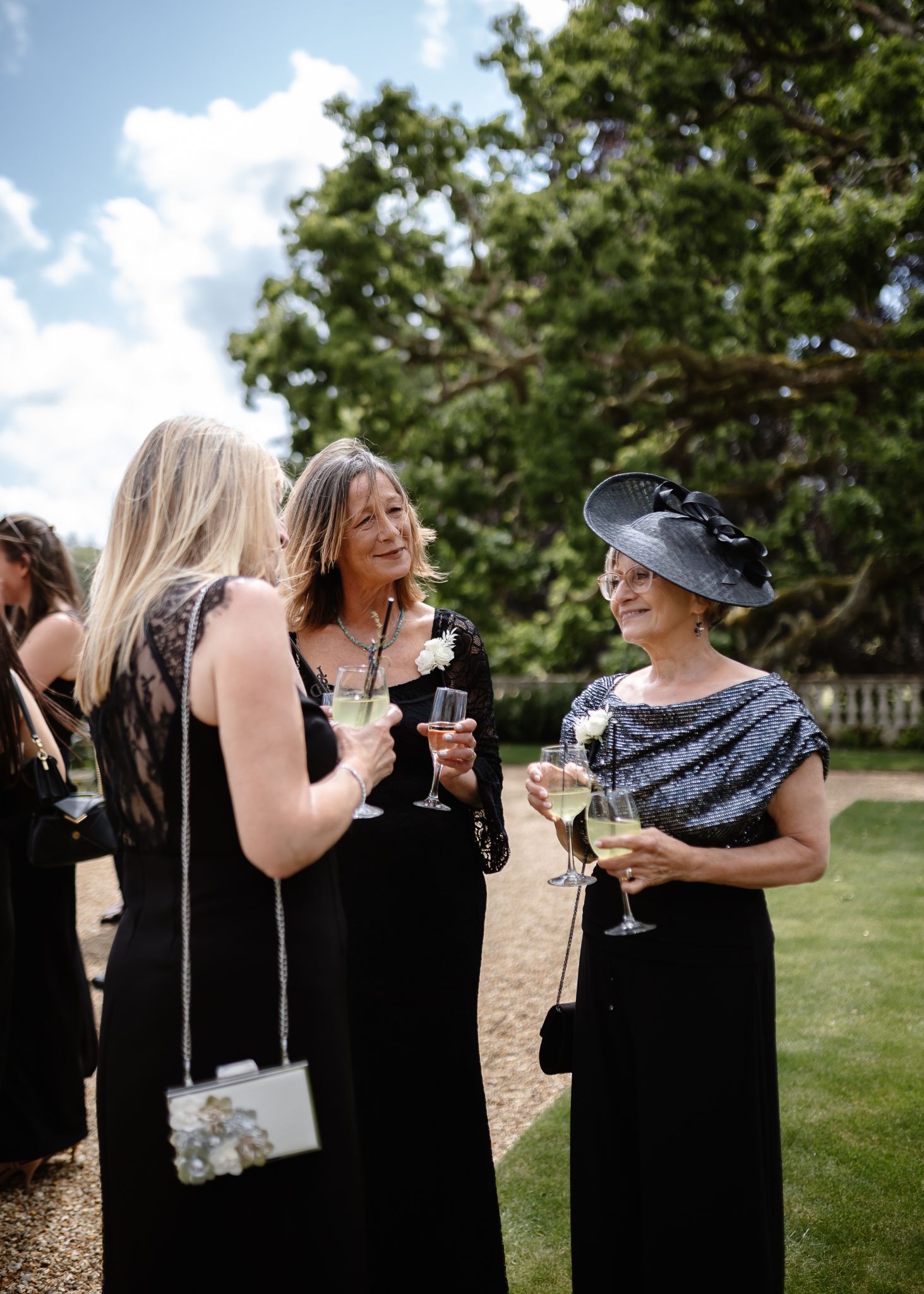 Wedding guests in black dresses socializing and laughing together at an outdoor celebration.