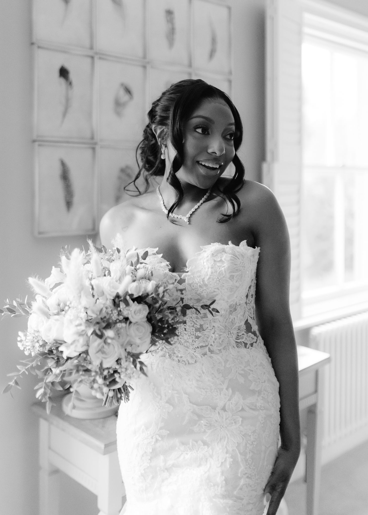 Black and white photo of a smiling bride in a strapless lace gown holding a bouquet, standing by a window.