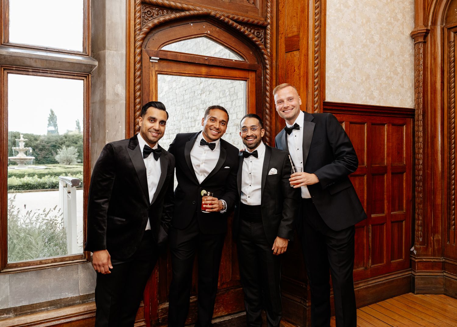 A group in formal black tuxedos and bow ties gather near a wood-paneled wall for a celebratory photo.