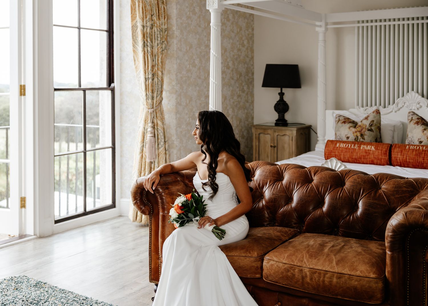 Elegant wedding dress displayed in a bright modern room with brown leather sofa and white curtains.