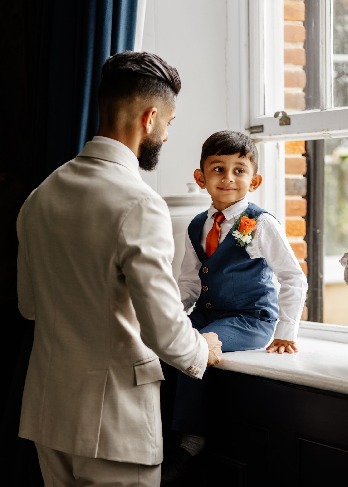 An adult and child in formal attire share a tender moment by a sunlit window.