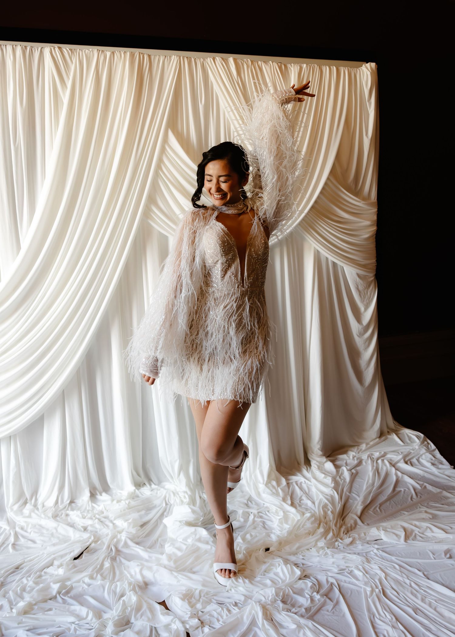 Woman in feathered white mini dress poses joyfully against draped ivory fabric backdrop.
