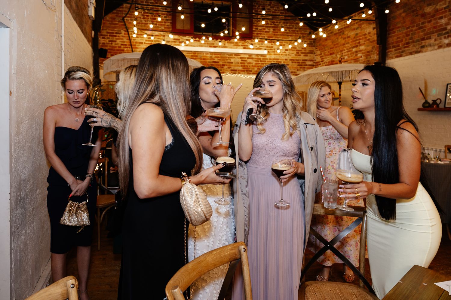 Wedding guests dance and celebrate at a rustic reception venue with exposed brick walls and twinkling lights.