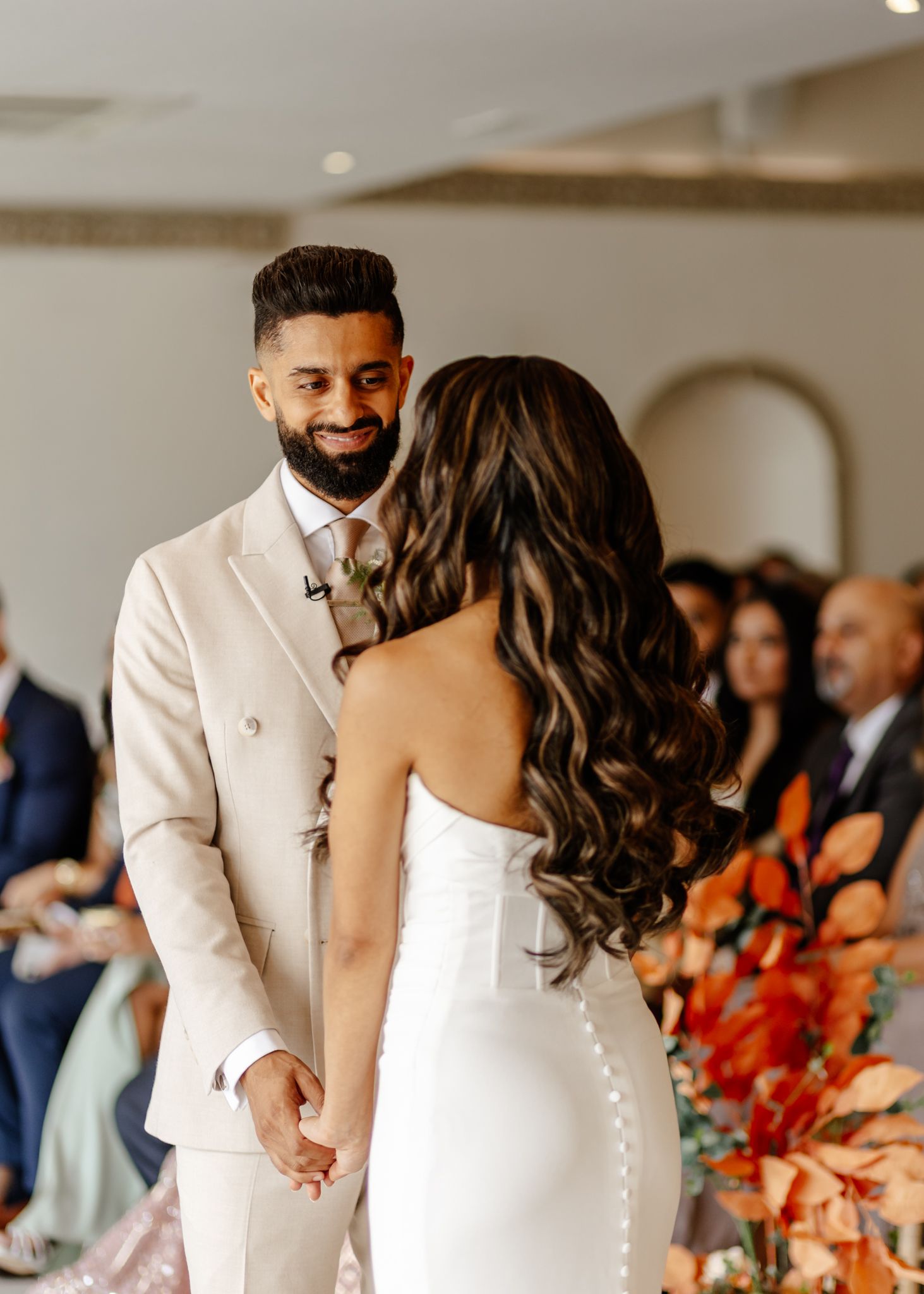 A couple shares a tender moment during their wedding ceremony, with the bride in a fitted white gown and groom in cream suit.