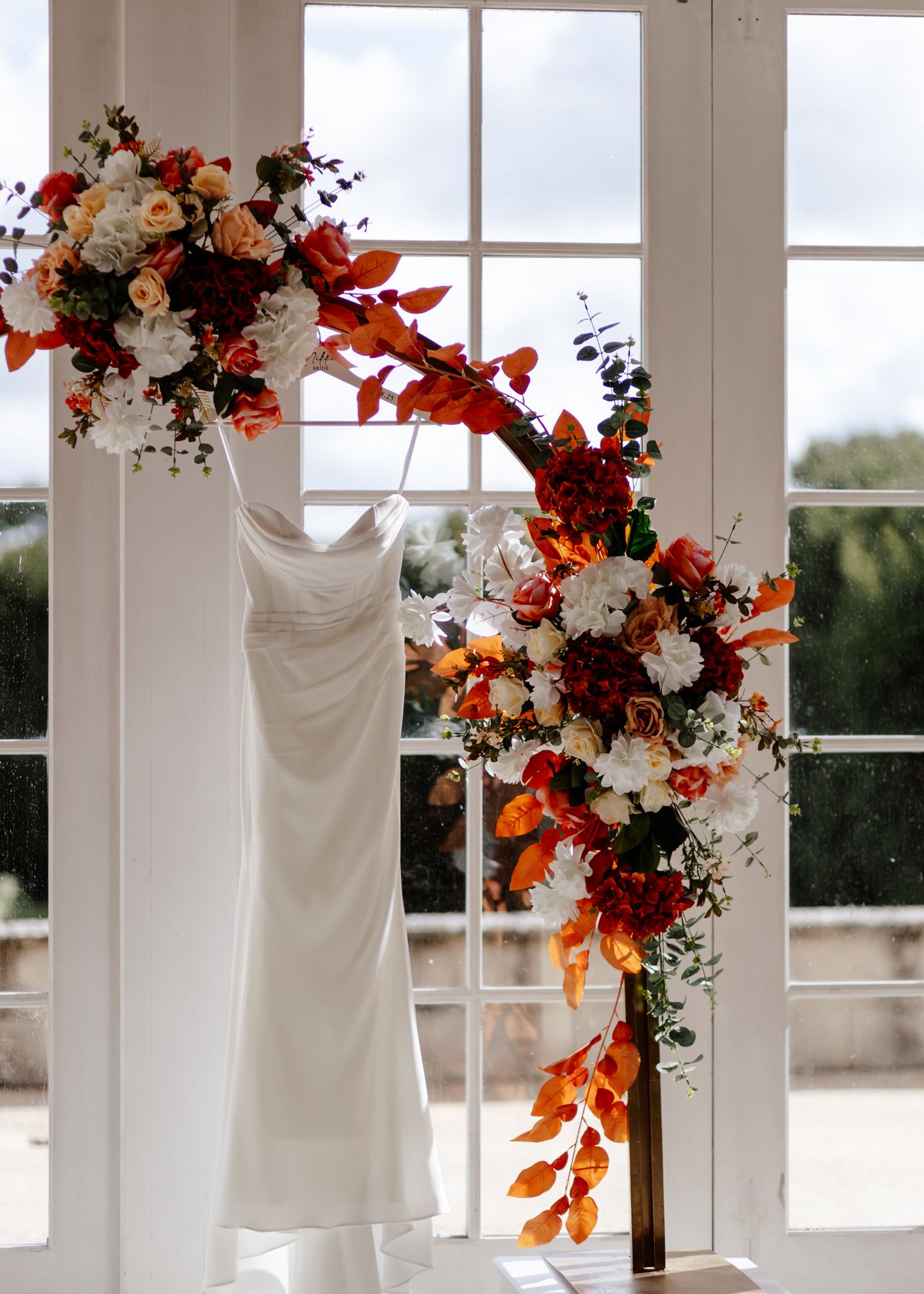 White wedding dresses hang in a bright room with autumn-colored floral arrangements decorating the windows.