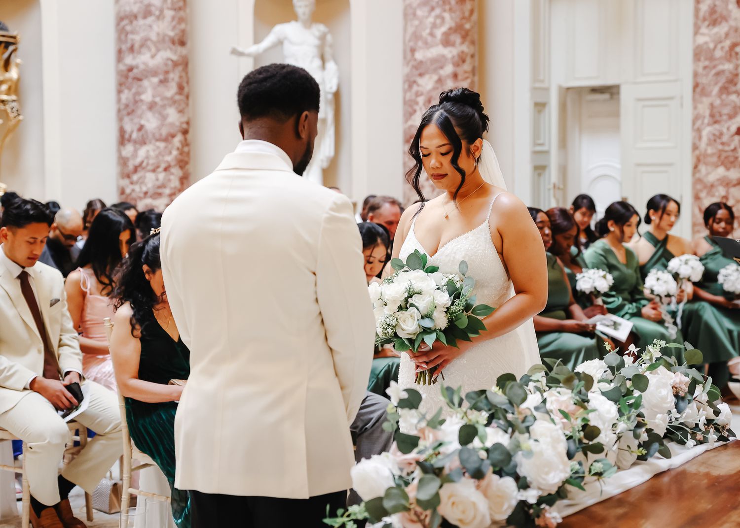 A wedding ceremony takes place in an elegant marble hall with pink columns and floral decorations.