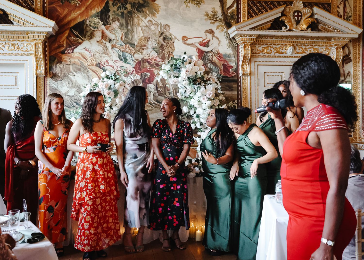 Wedding party members in colorful dresses stand in line against an ornate baroque interior with gilded details.