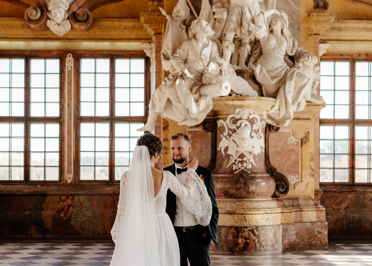 Elegant wedding couple dances in a historic baroque palace hall with ornate architectural details and large windows.