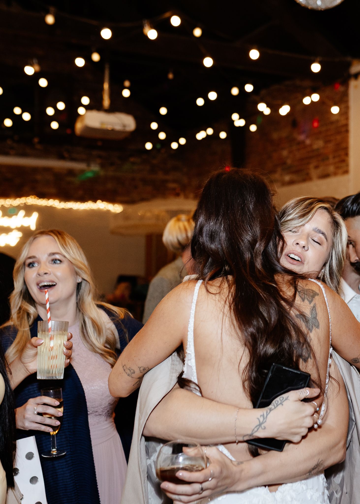People dancing and celebrating at a wedding reception with string lights overhead and drinks in hand.
