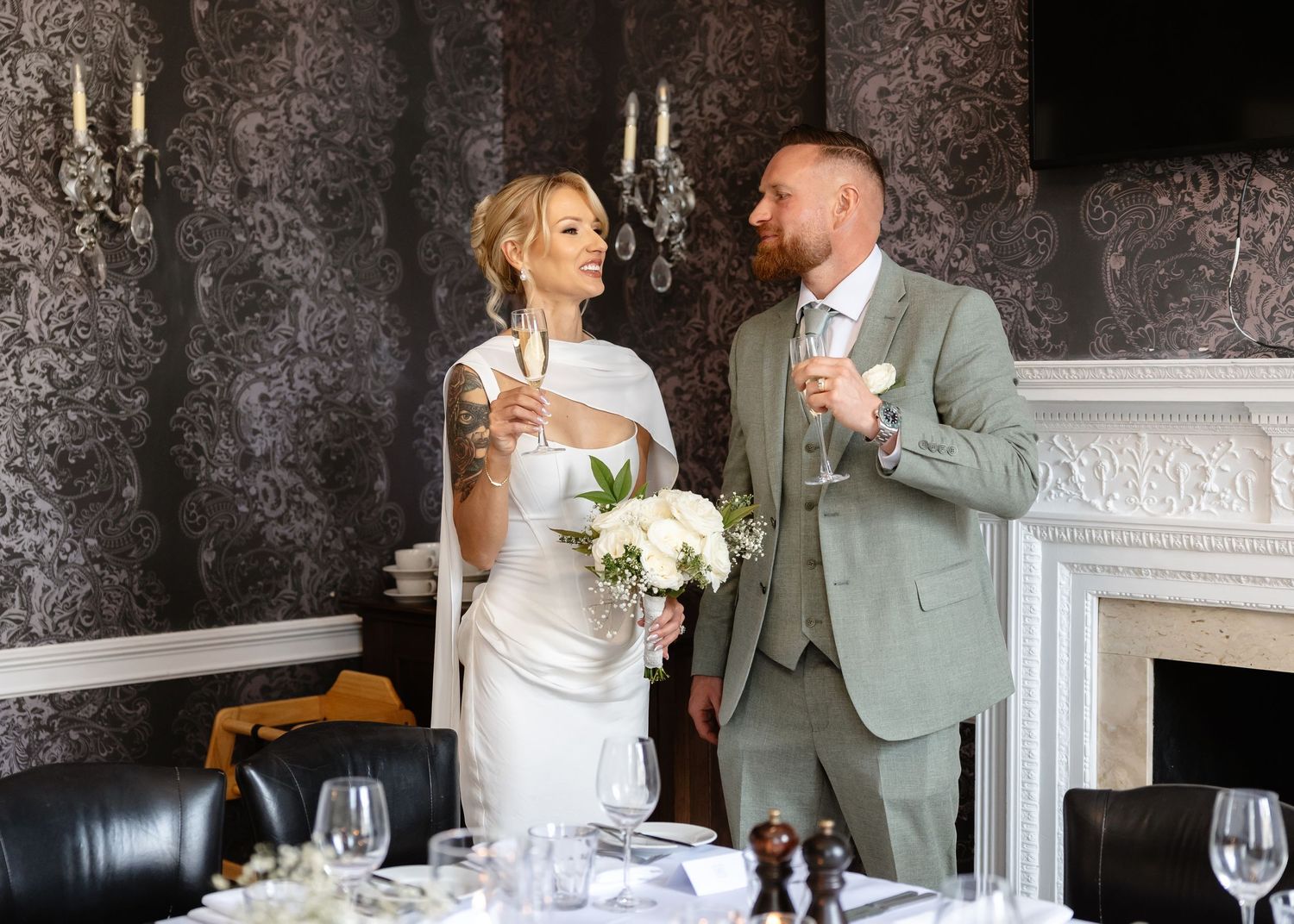 Bride and groom toasting with champagne at wedding reception, bride holding white bouquet, groom in sage green suit.