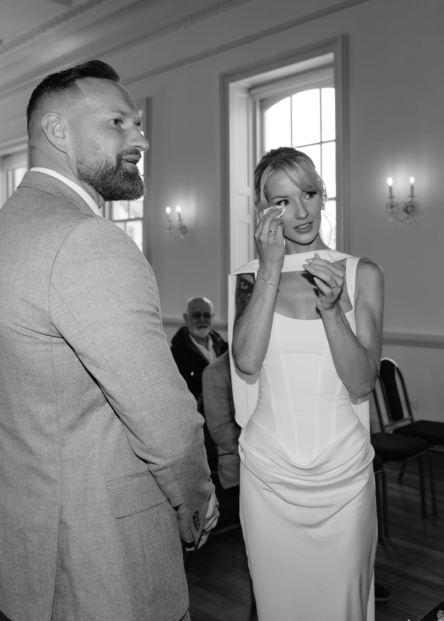 Emotional bride wiping tears during wedding ceremony while groom stands beside her in elegant venue.