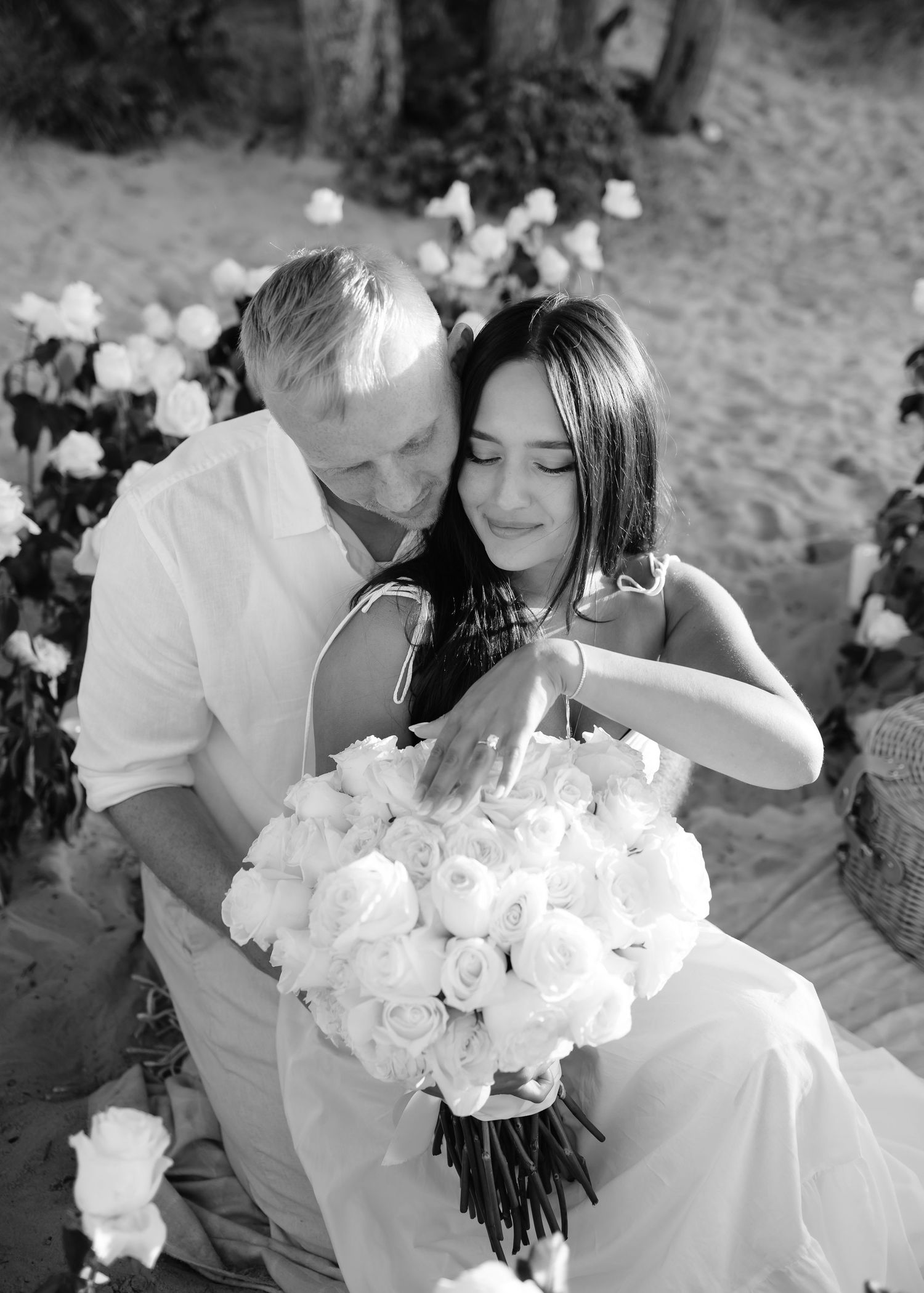 Black and white photo of a couple embracing on a beach, woman holding a large white rose bouquet, surrounded by flowers.