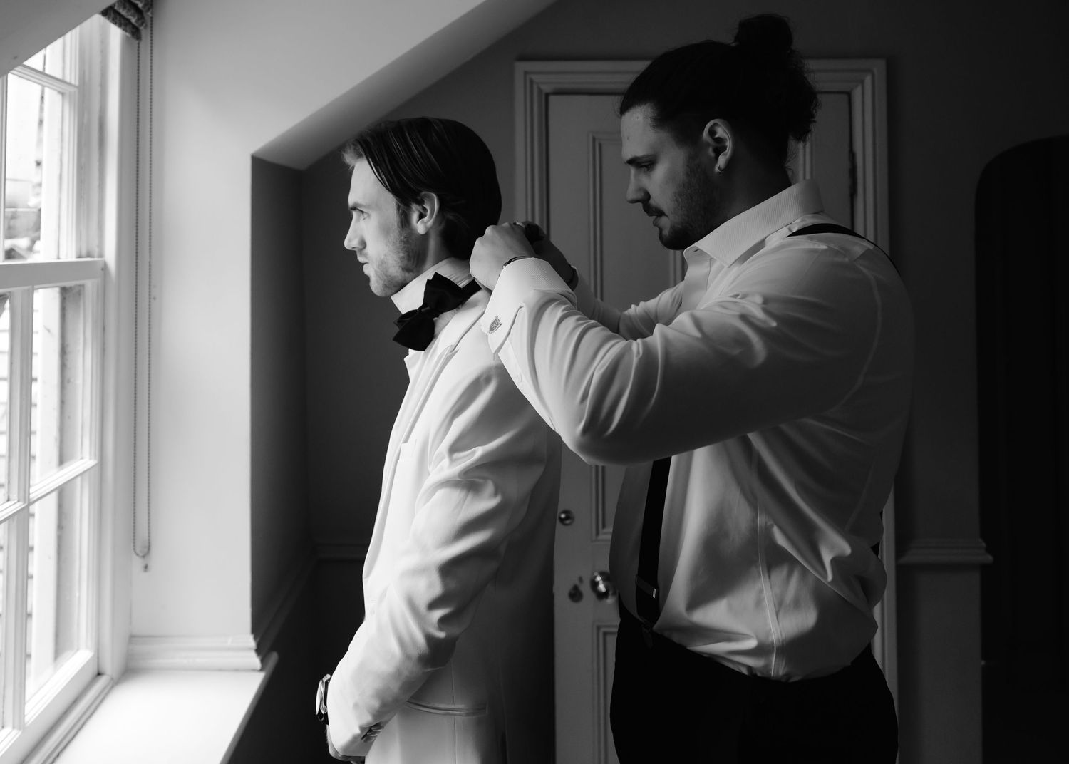 Black and white photo of a groomsman helping a groom adjust his bow tie before a wedding ceremony.