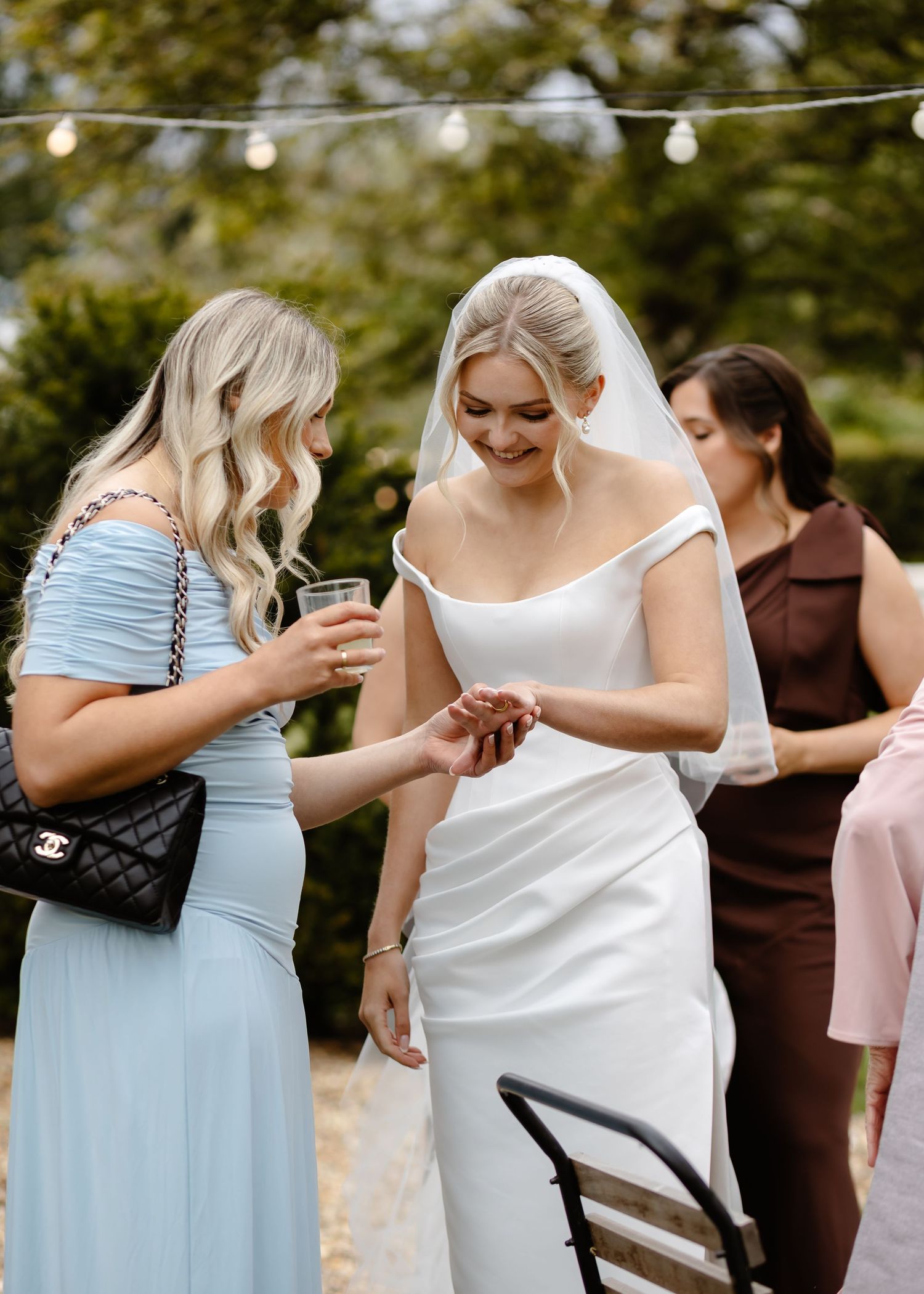 Bride in white gown shows her ring to a bridesmaid in light blue dress at an outdoor wedding reception.