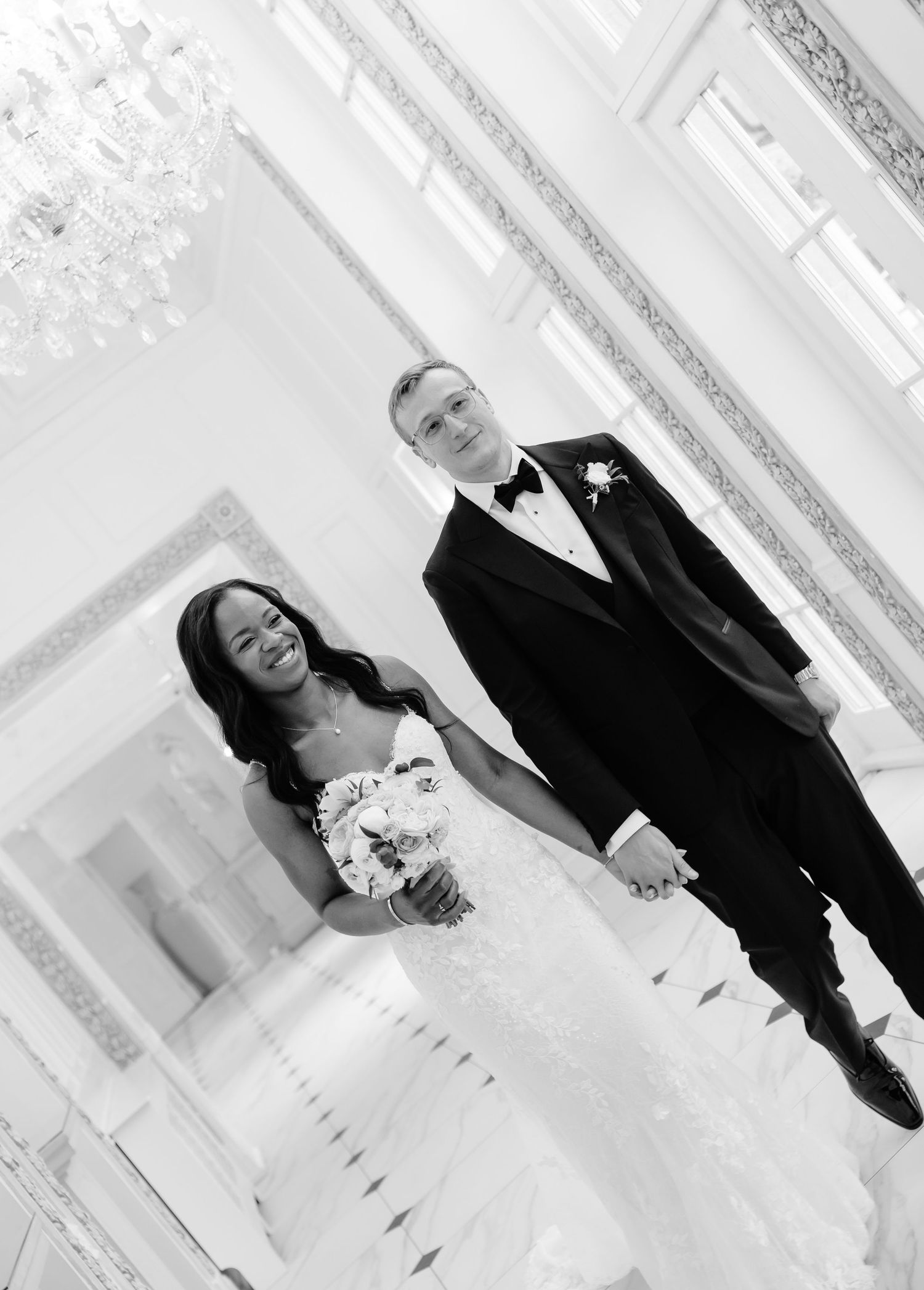 Black and white wedding photo of smiling bride in strapless gown holding bouquet standing with groom in tuxedo in elegant venue.
