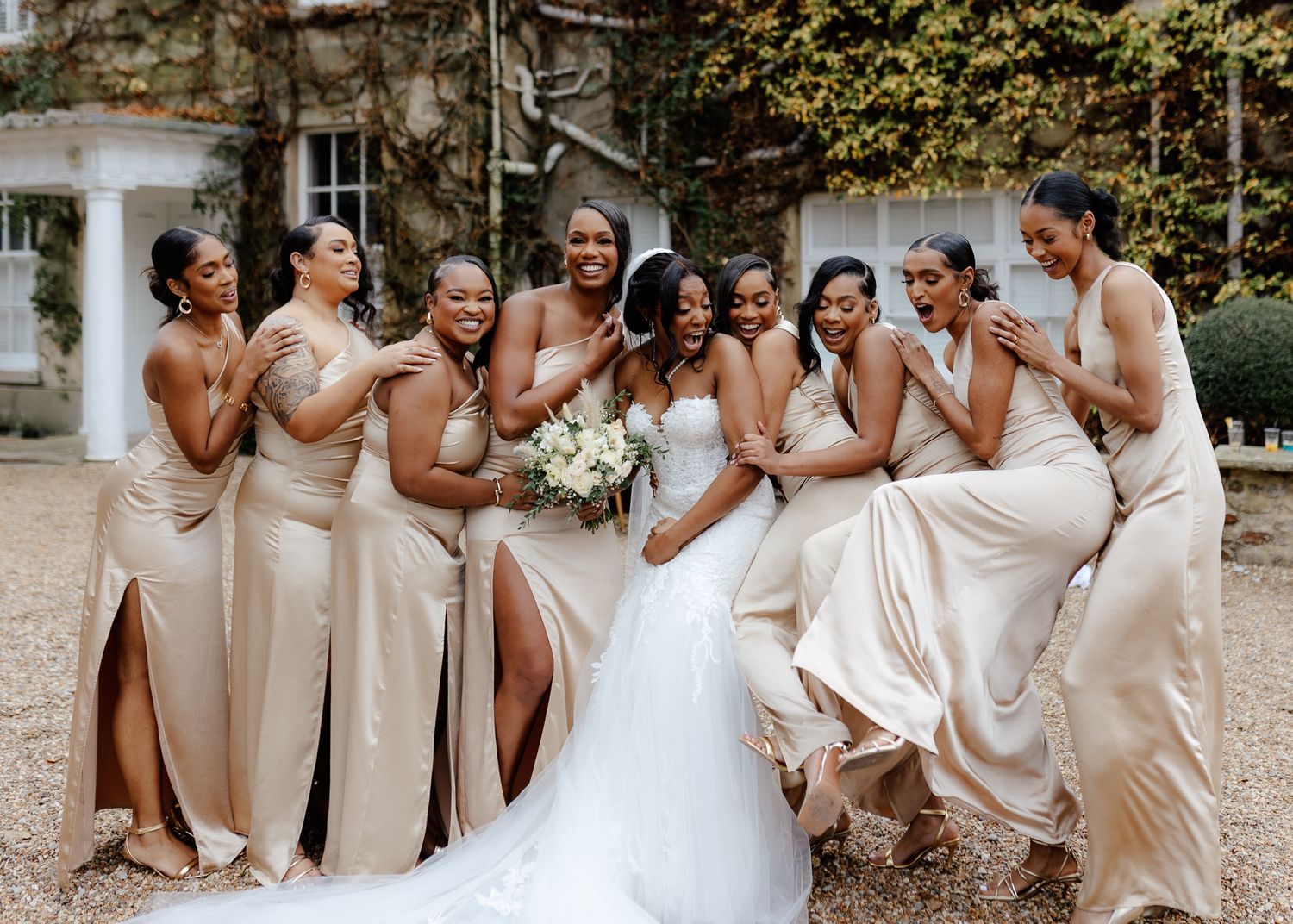Bridal party in champagne-colored dresses share joyful laughter during outdoor wedding photo session.