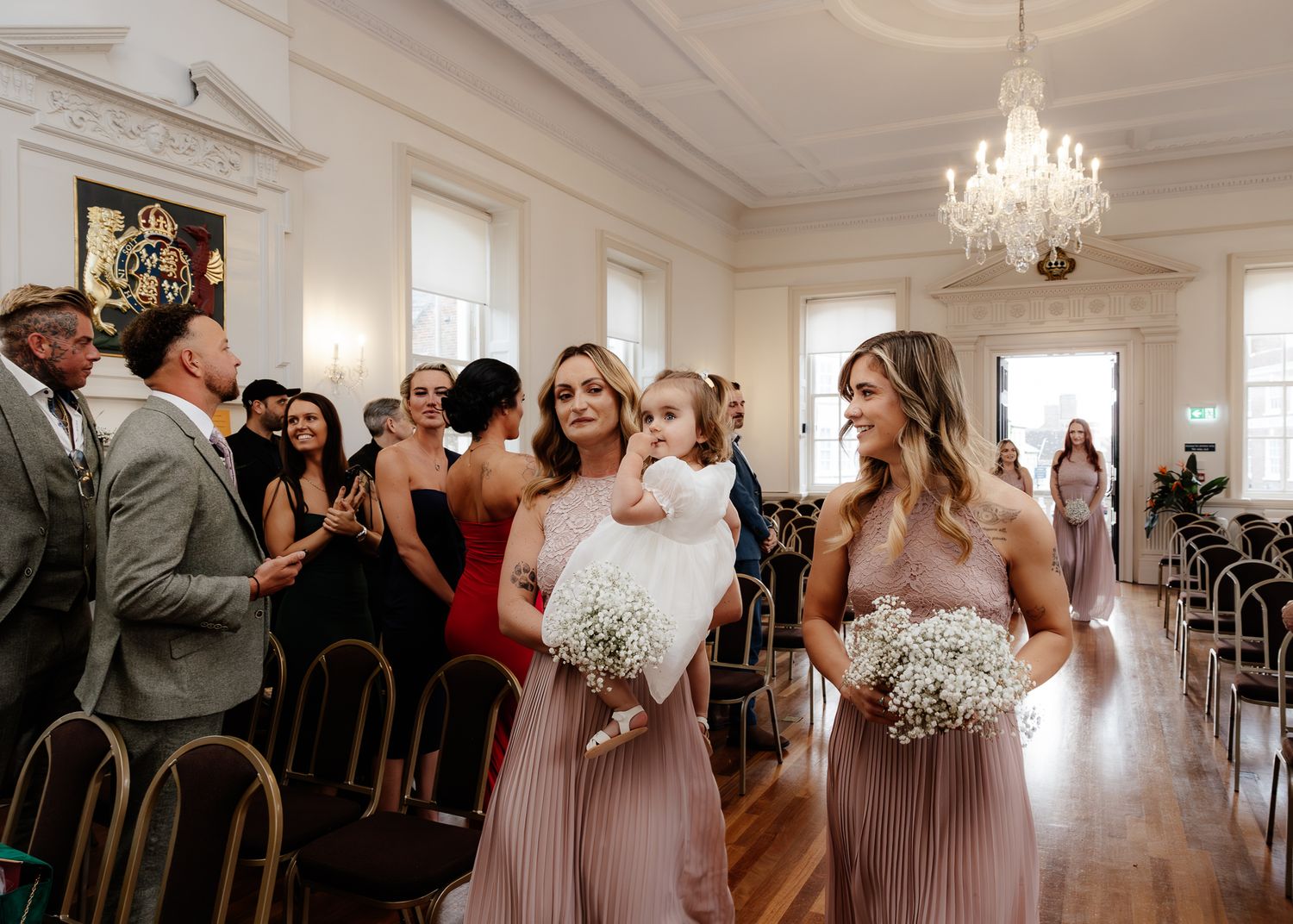 Bridesmaids in pink dresses hold white bouquets while standing in an elegant room with a crystal chandelier.