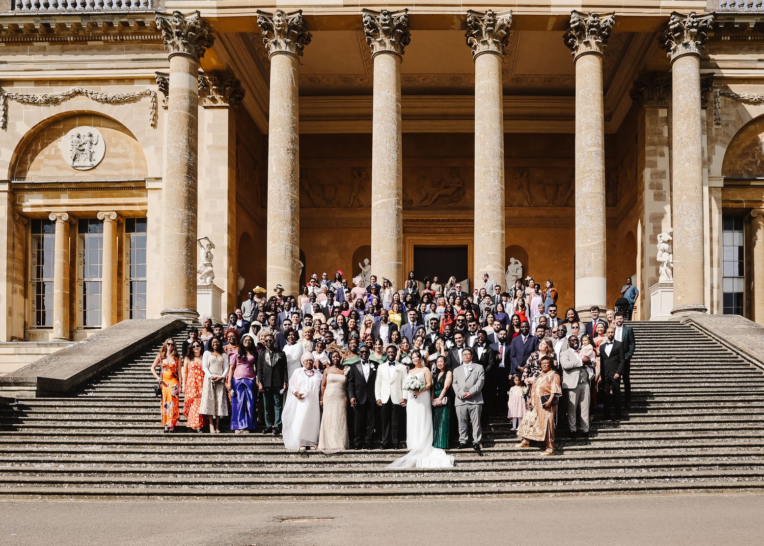 Large wedding party gathered on grand stone steps of historic building with classical columns for panoramic group photo.