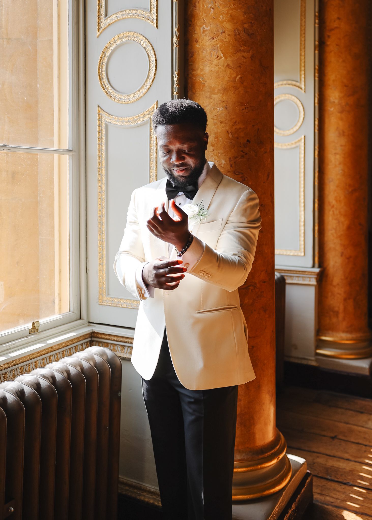 Person in a white blazer and black pants stands in an elegant room with ornate columns and architectural details.