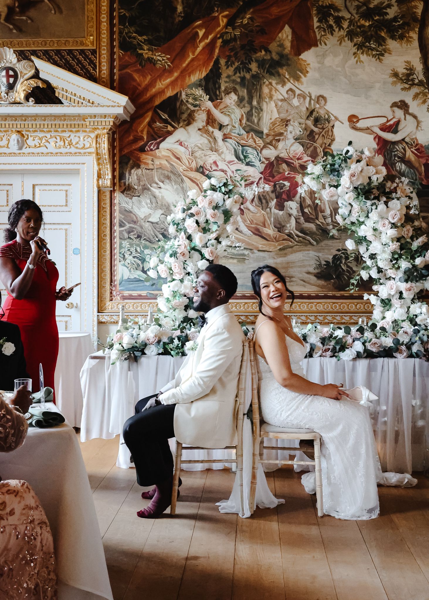 Wedding party members dance and celebrate in an elegant ballroom decorated with white floral arrangements and ornate decor.