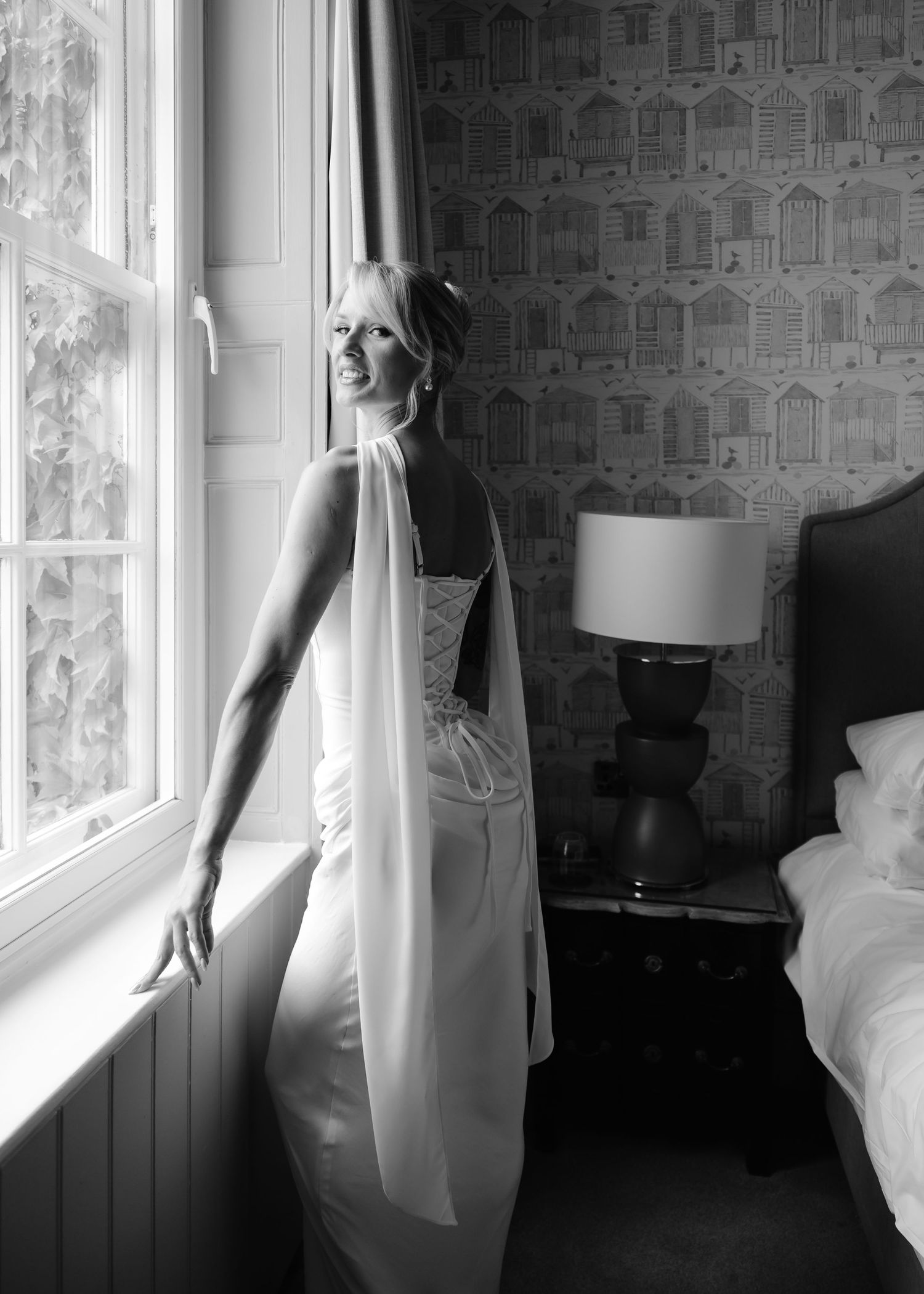 Bride in white wedding gown with veil stands by window in elegant bedroom, black and white photography.