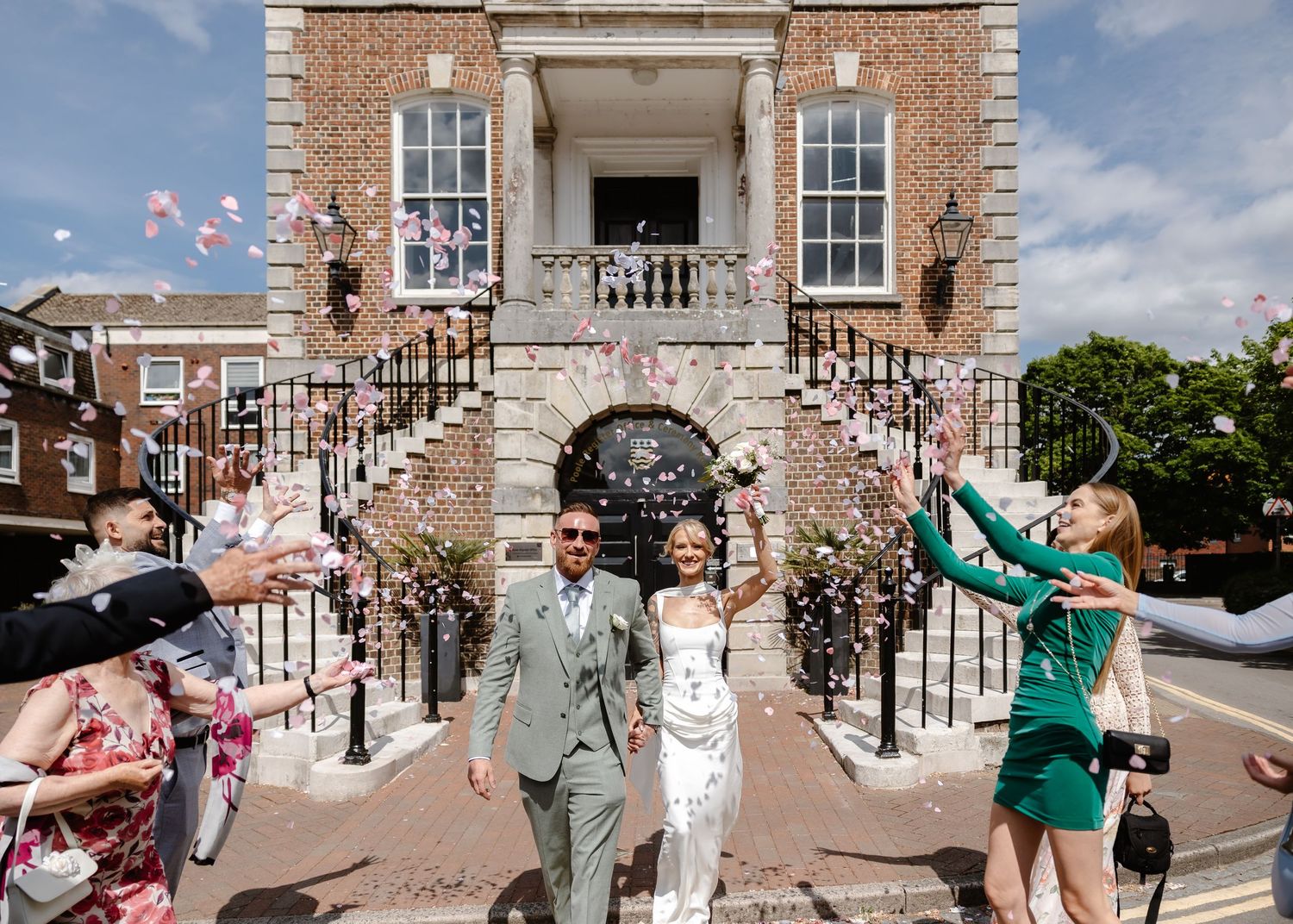 Bride and groom exit a brick building showered with confetti by guests on a sunny wedding day.