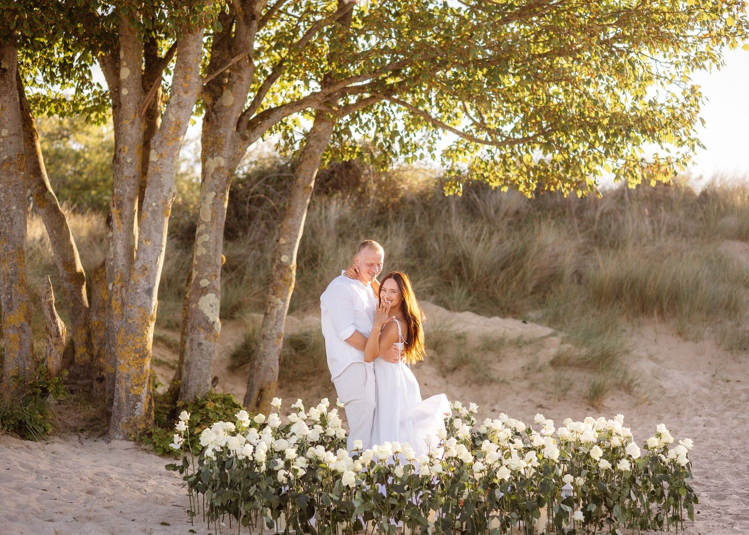 Couple in white embracing on sandy beach surrounded by white flowers and trees in golden hour light.
