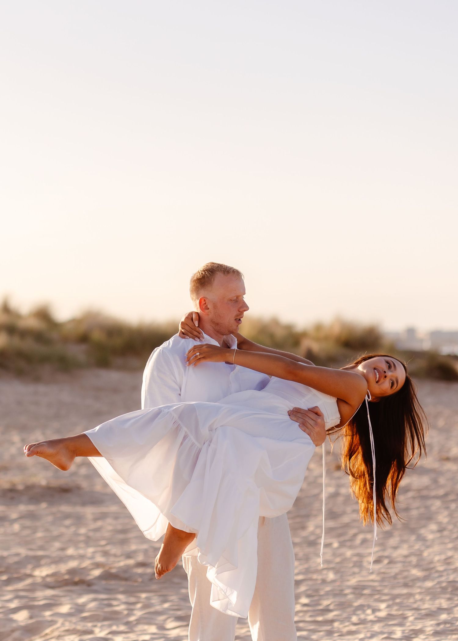 Man in white shirt lifting woman in white dress on sandy beach at sunset.