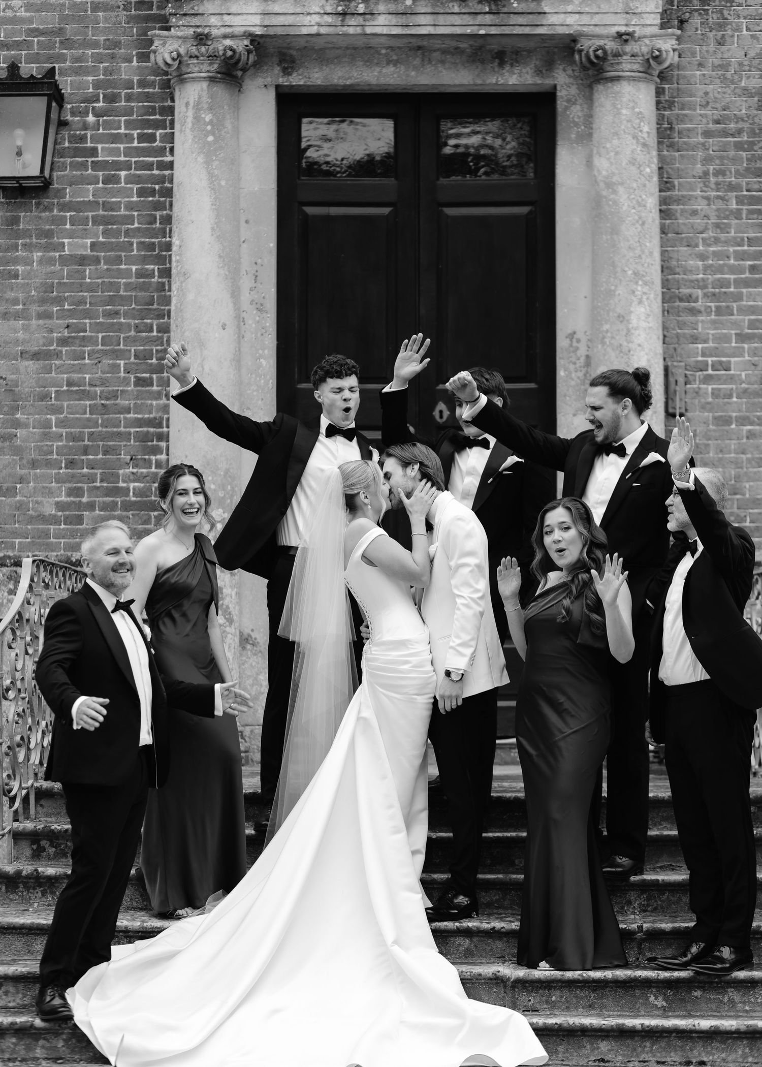 Black and white wedding photo of couple kissing on steps surrounded by cheering wedding party in formal attire.