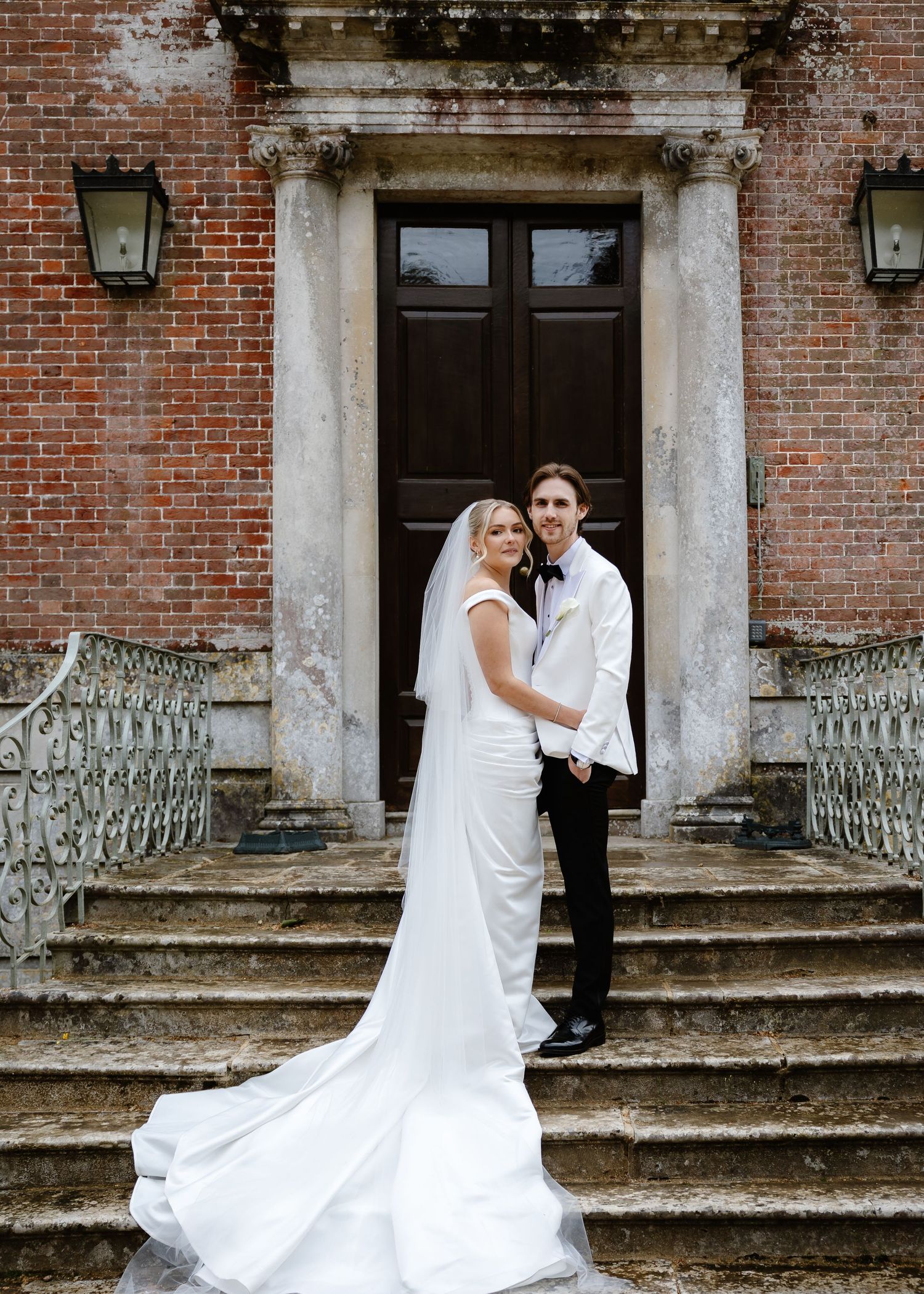 Bride and groom pose on stone steps before a brick manor house, bride in elegant white gown with long veil.