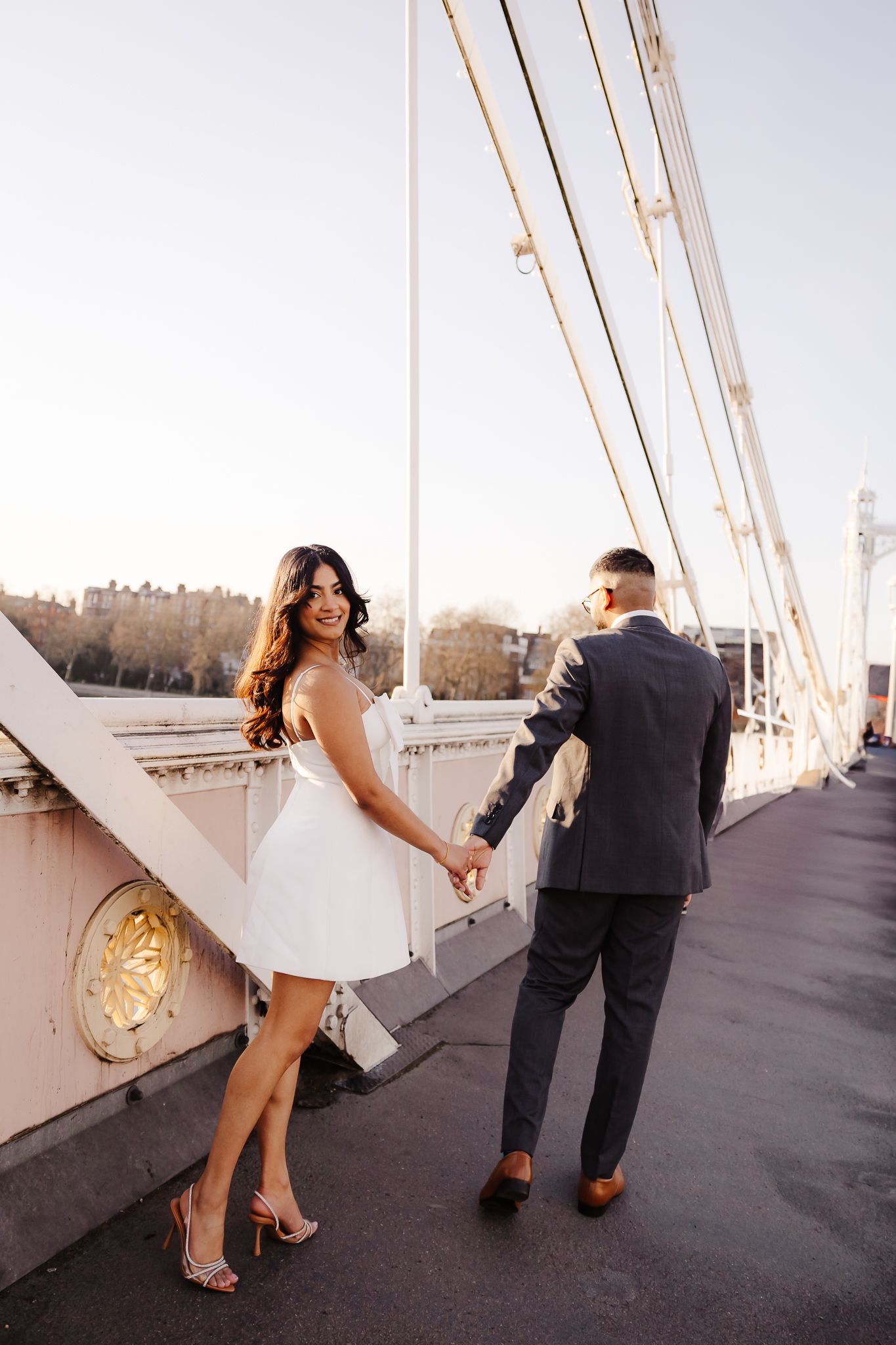 Bride in white mini dress and groom in navy suit hold hands on a sunlit suspension bridge.