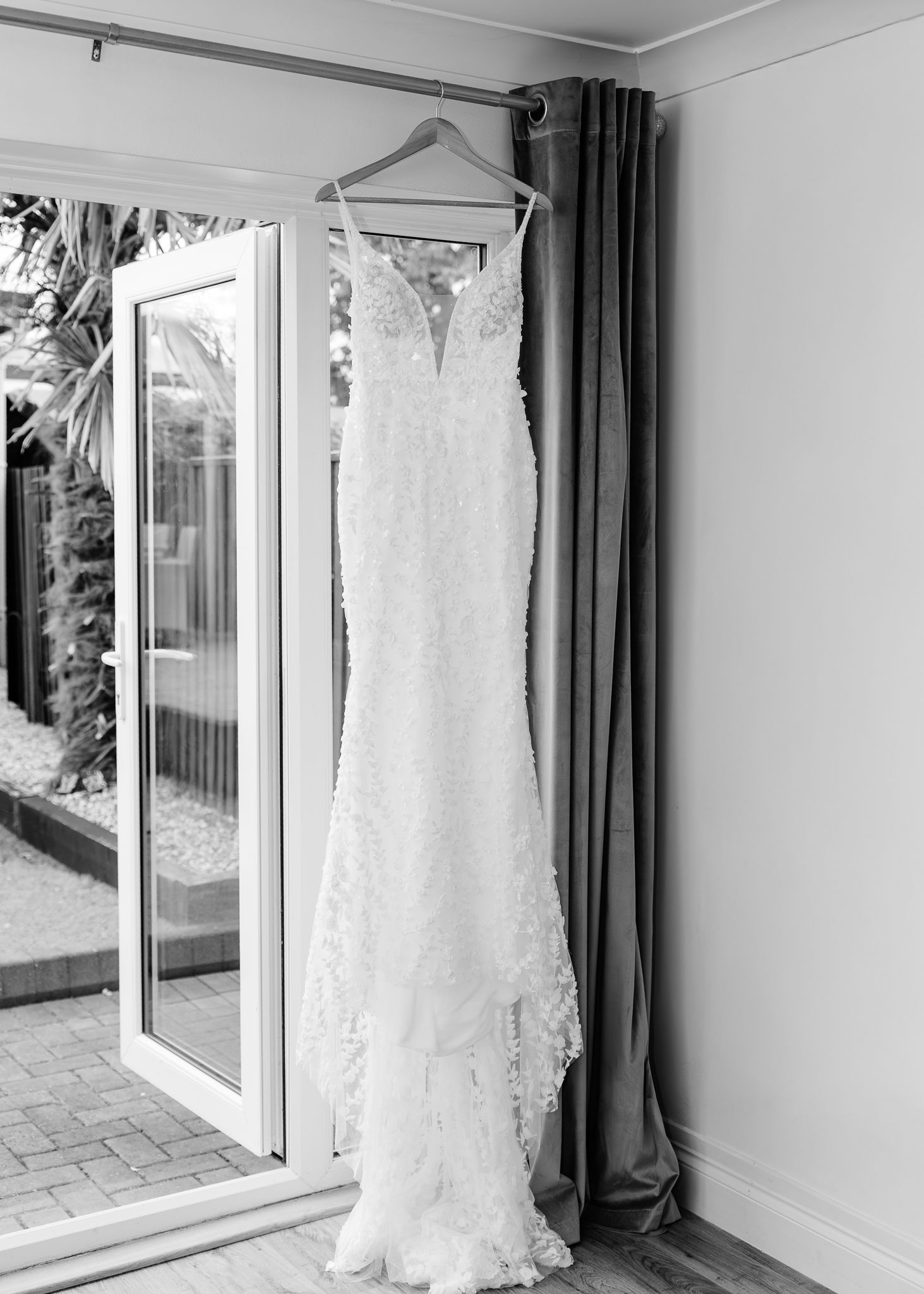 White lace wedding dress hanging on display in a bright room with large windows and dark curtains.