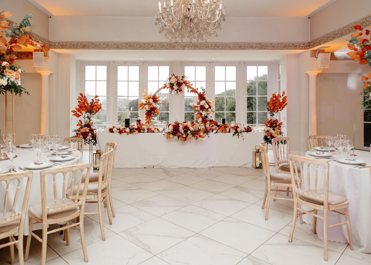 Elegant white wedding reception venue with chandeliers, white chairs, and autumn floral decorations.