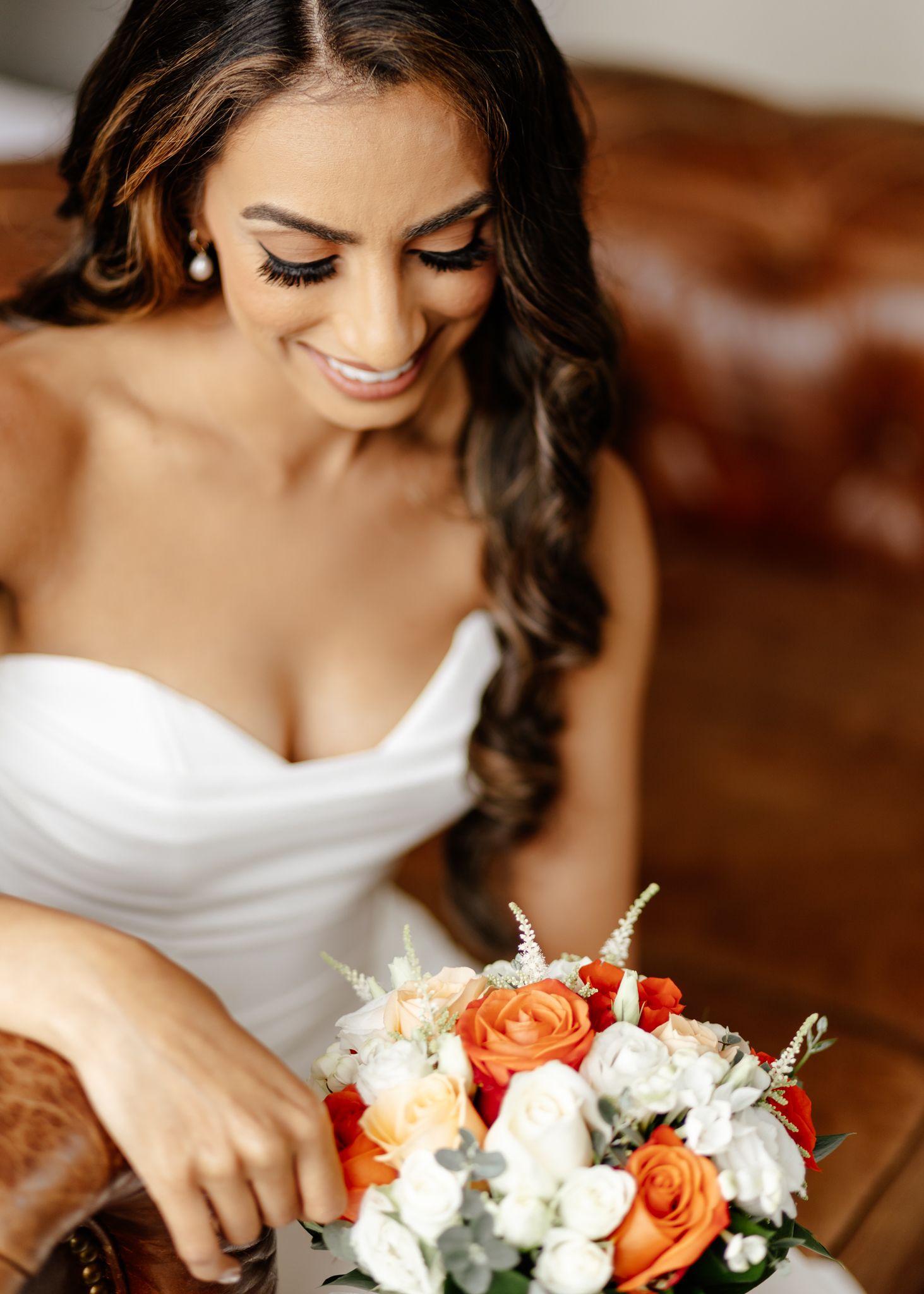 Bride in white strapless dress holding a vibrant orange and white rose bouquet during wedding preparations.
