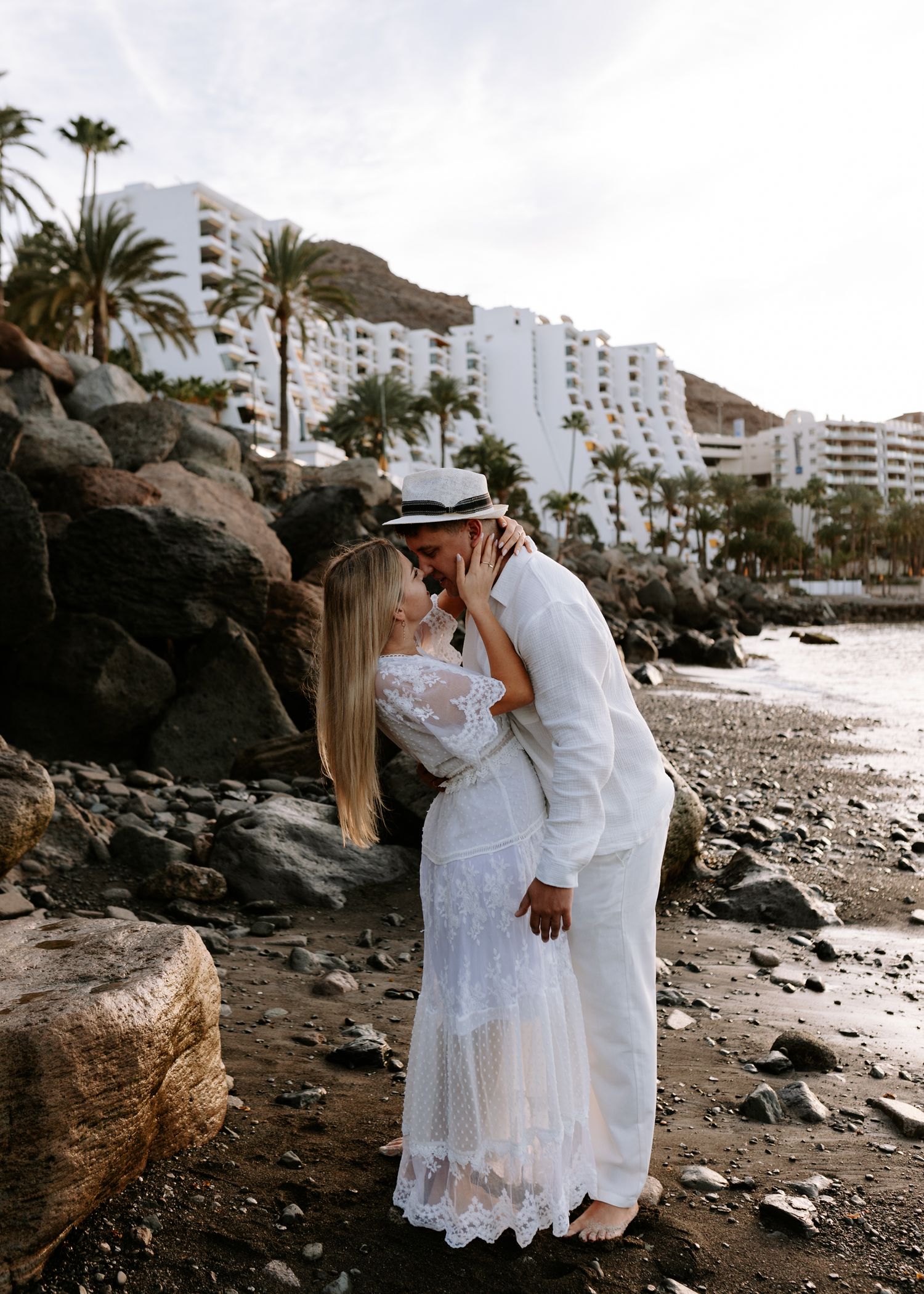 A couple in white outfits share romantic moments on a rocky beach with palm trees and buildings in the background.