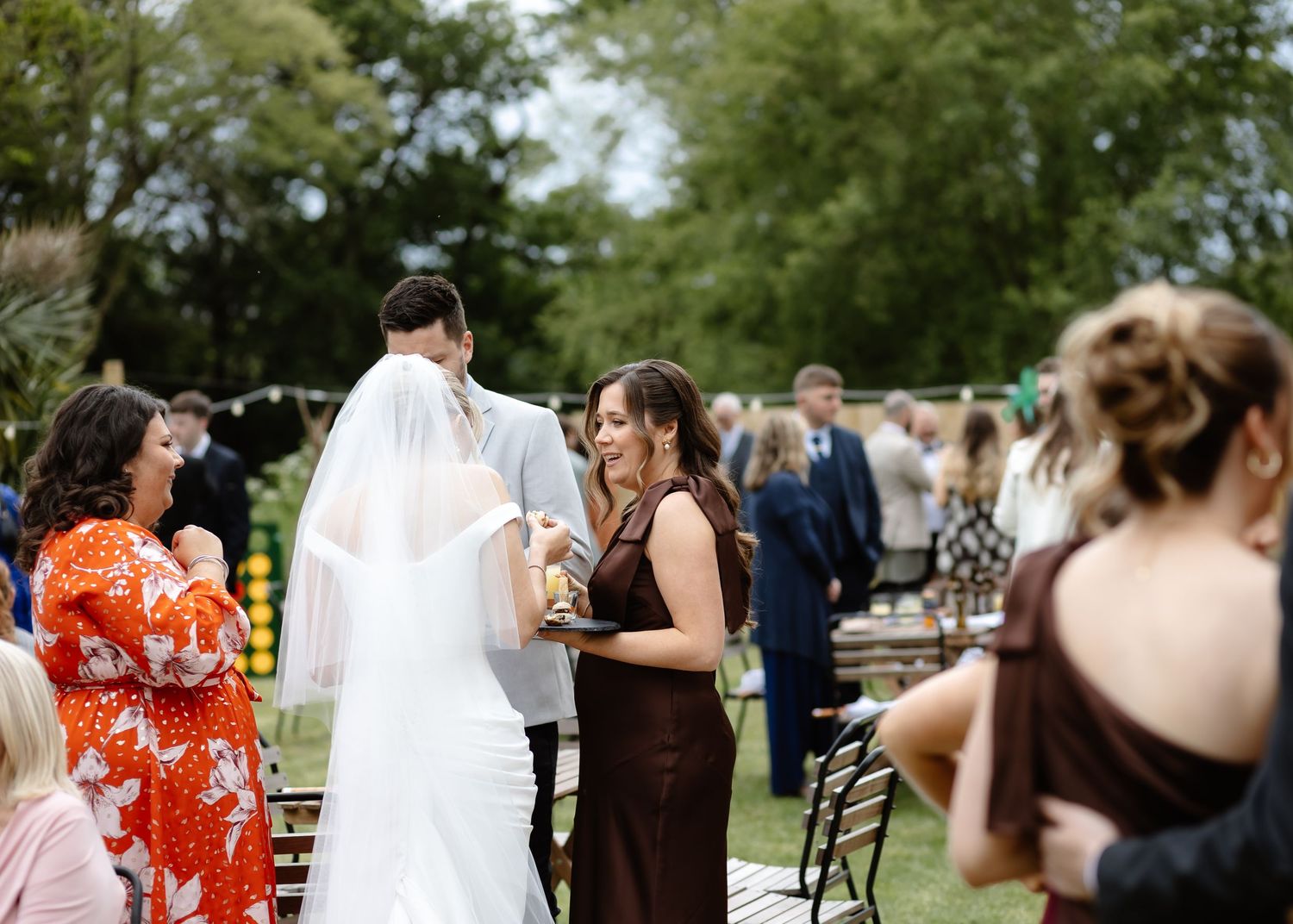 Outdoor wedding ceremony with bride in white gown and veil, groom, and bridesmaid in brown dress among guests.