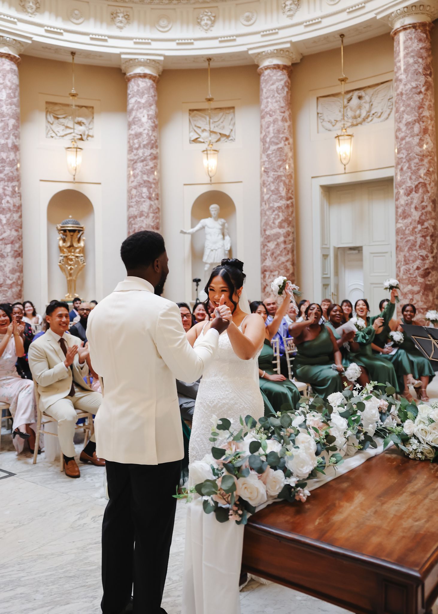 A wedding celebration in an elegant circular room with marble columns and floral decorations on wooden tables.