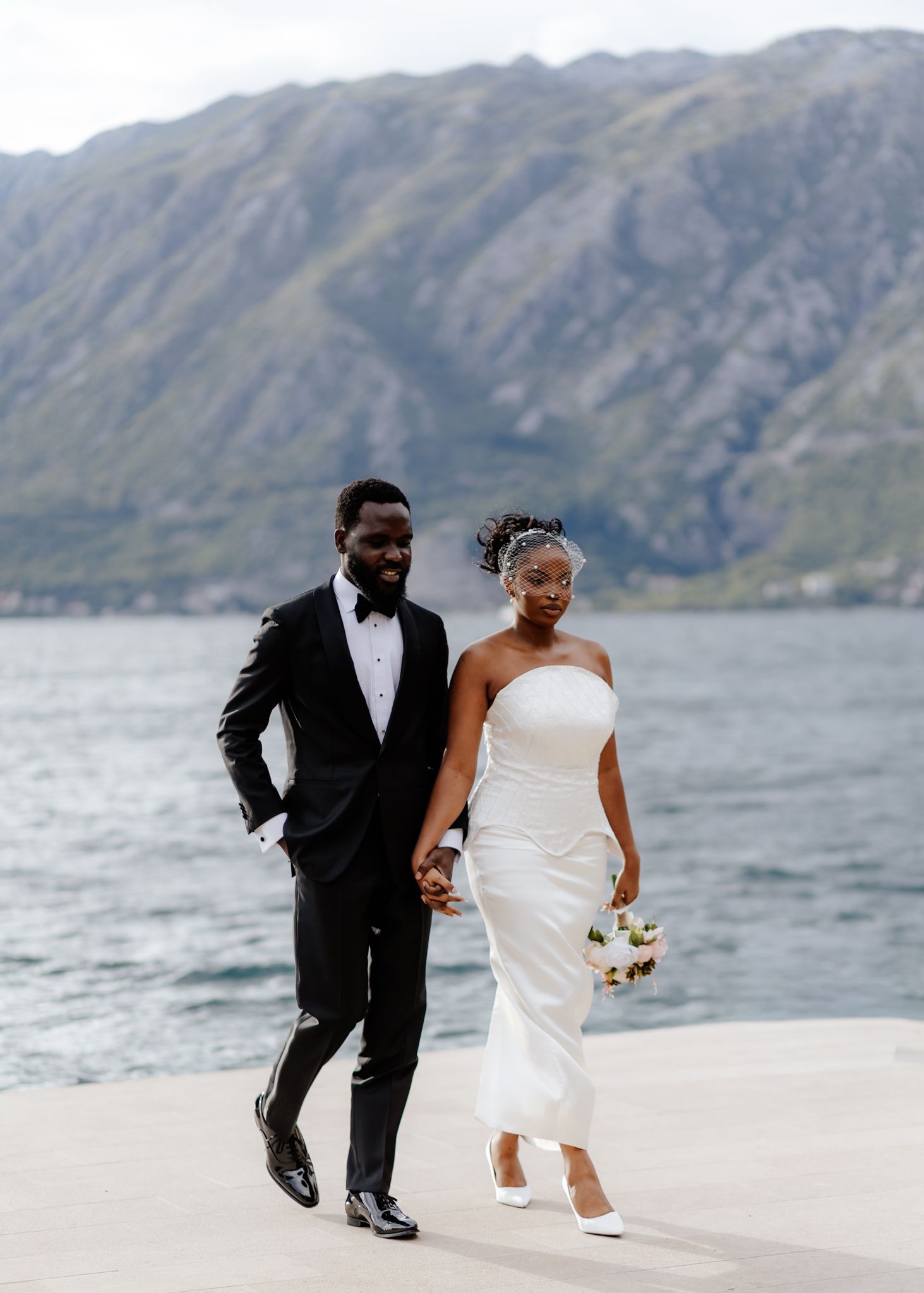 Newlywed couple runs playfully along a scenic beach with mountains and water in the background.