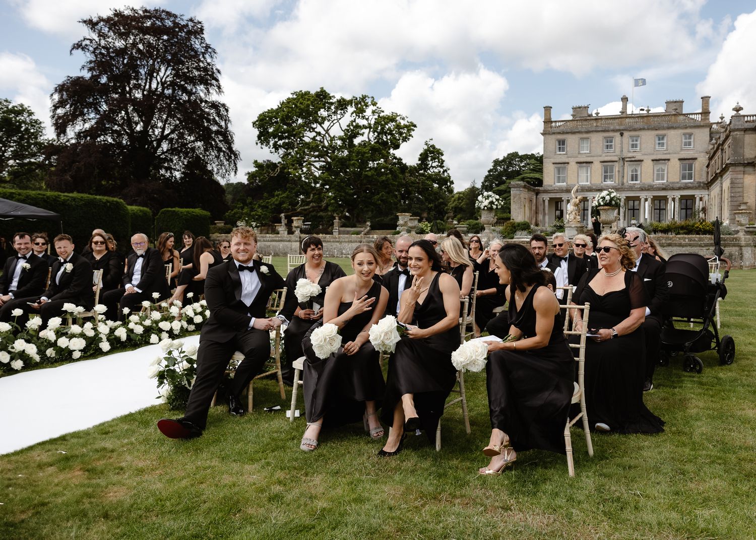 Wedding guests seated on a grassy lawn in front of a grand estate during an outdoor ceremony.