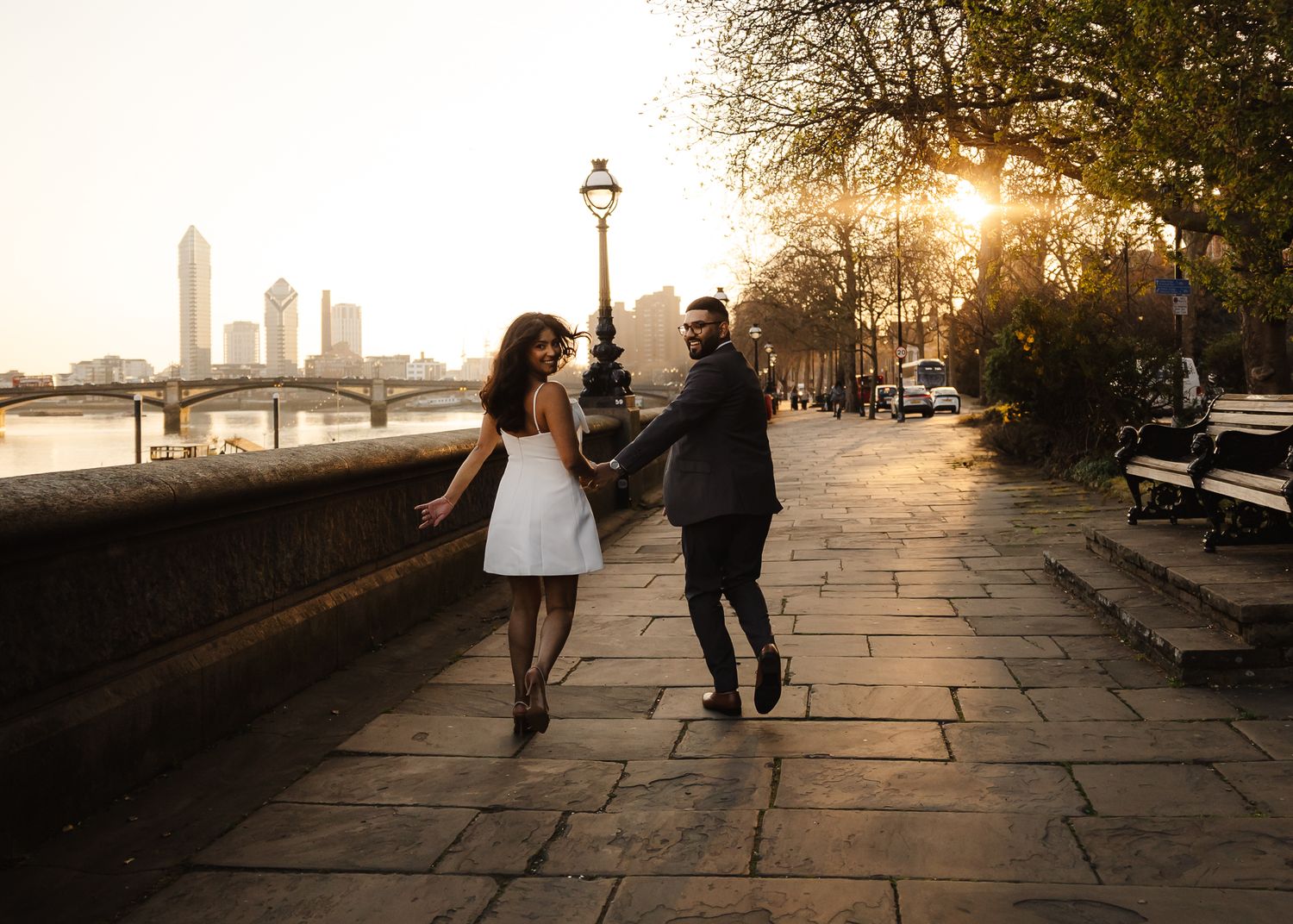 Silhouettes of people walking along a waterfront promenade at sunset with golden light streaming through trees.