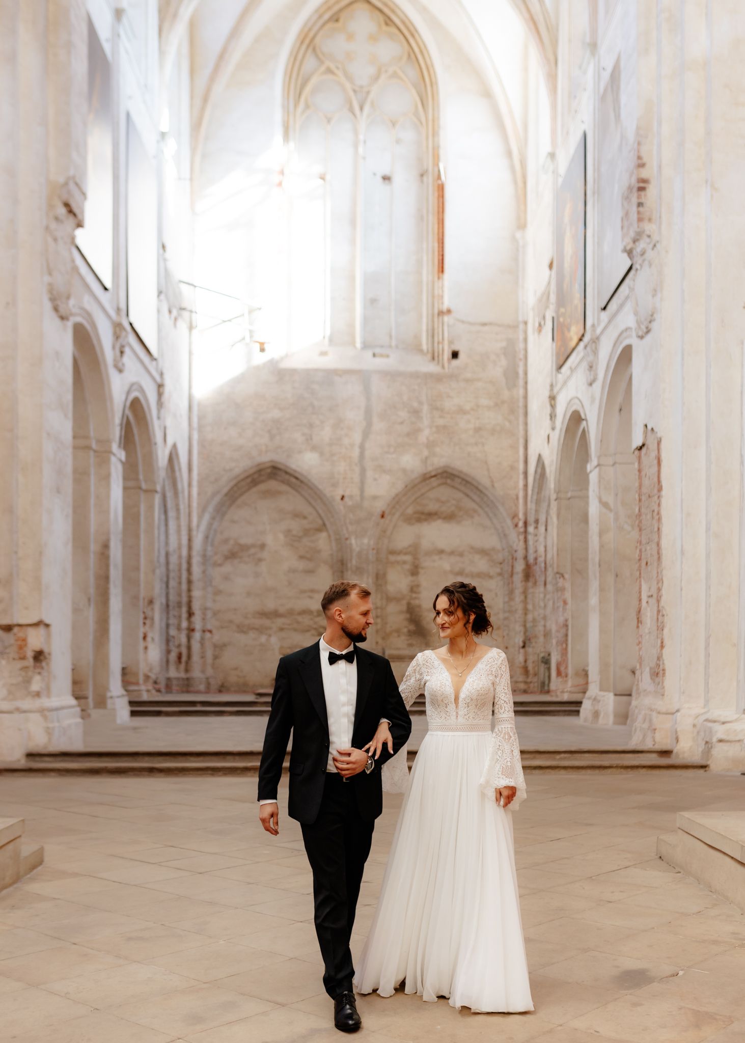 Wedding couple walking through a bright Gothic church with high vaulted ceilings and white walls.