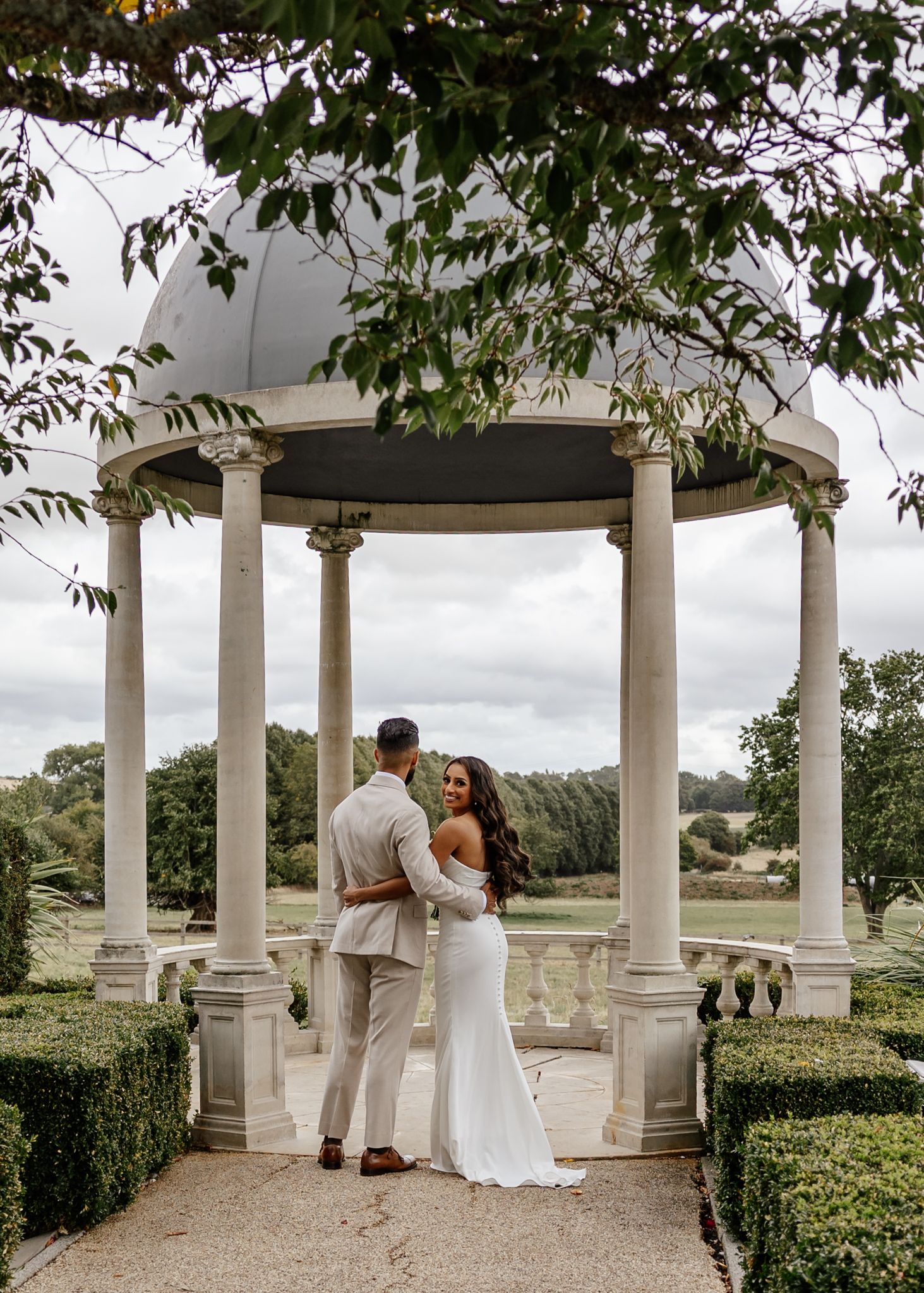 A couple shares a romantic moment under an elegant white garden gazebo with ornate columns and lush greenery overhead.