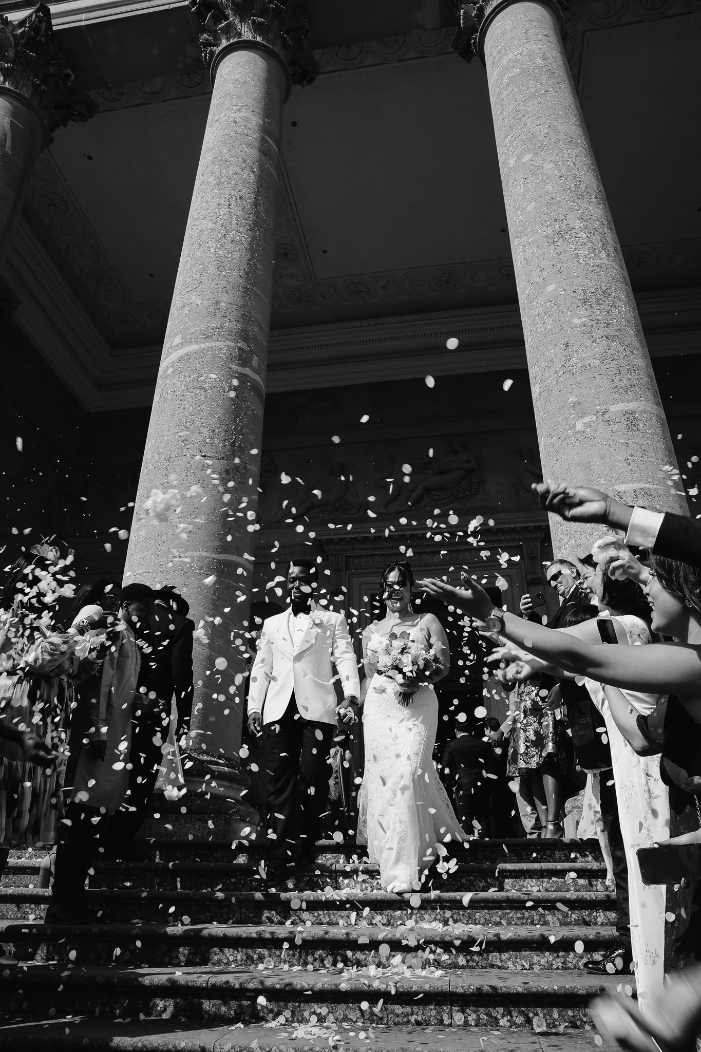 Black and white photo of wedding celebration with confetti being thrown between classical stone columns.