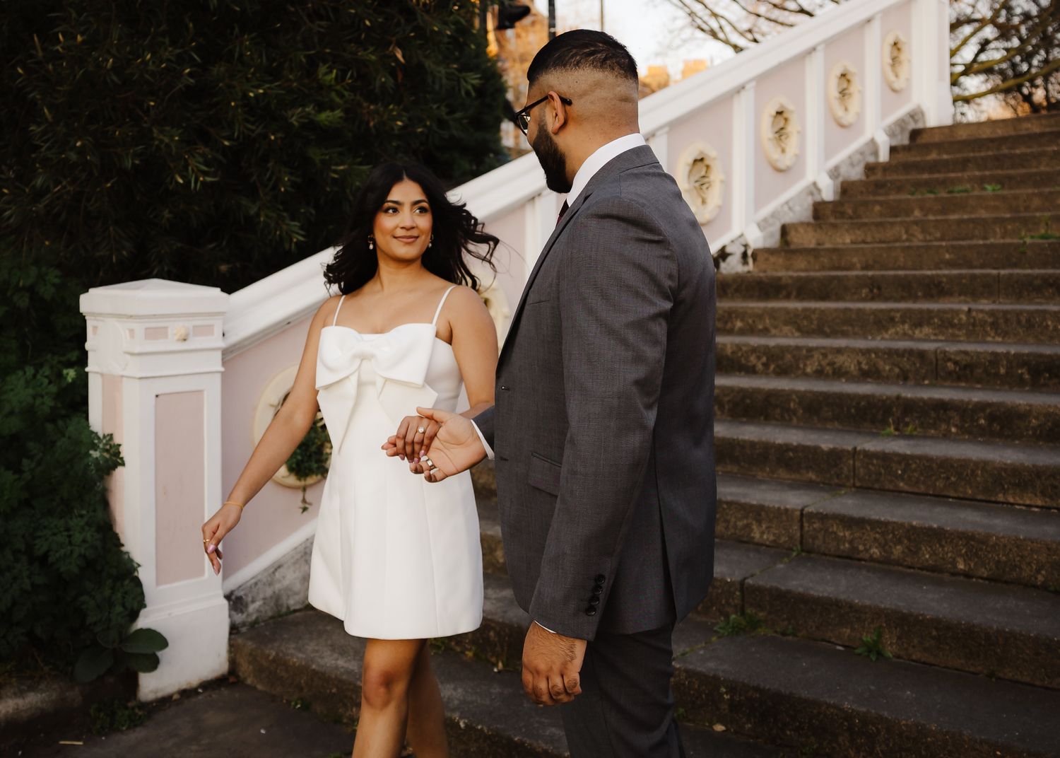 Couple holding hands near elegant white staircase, woman in white mini dress, man in gray suit, outdoor engagement photo.