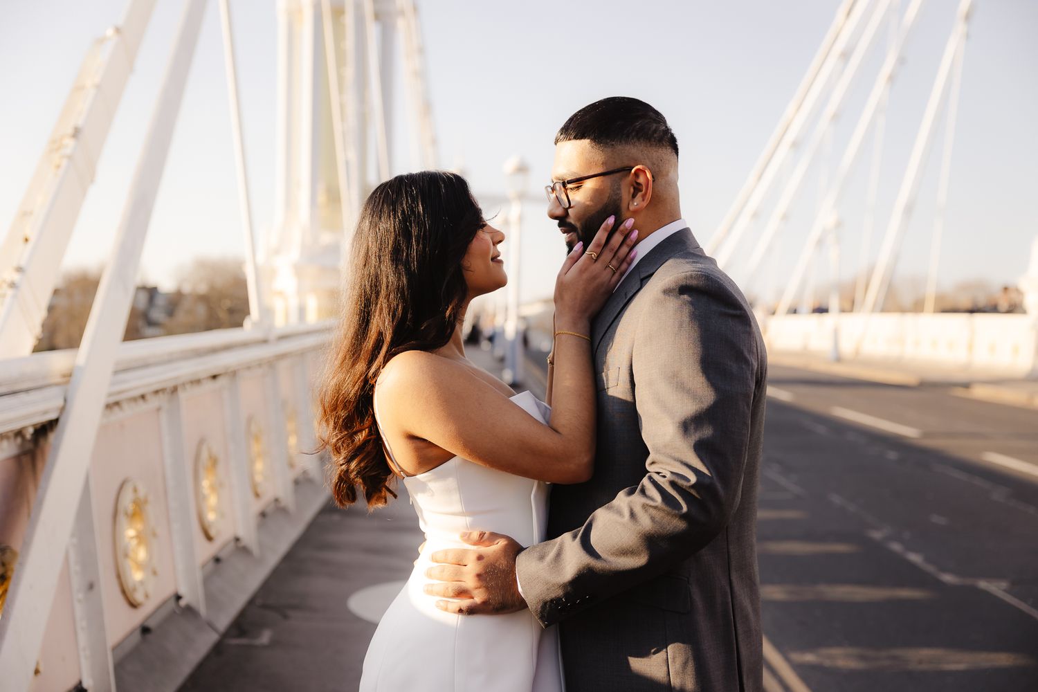 Couple sharing romantic moment on bridge, woman in white dress touching mans face, golden hour lighting.