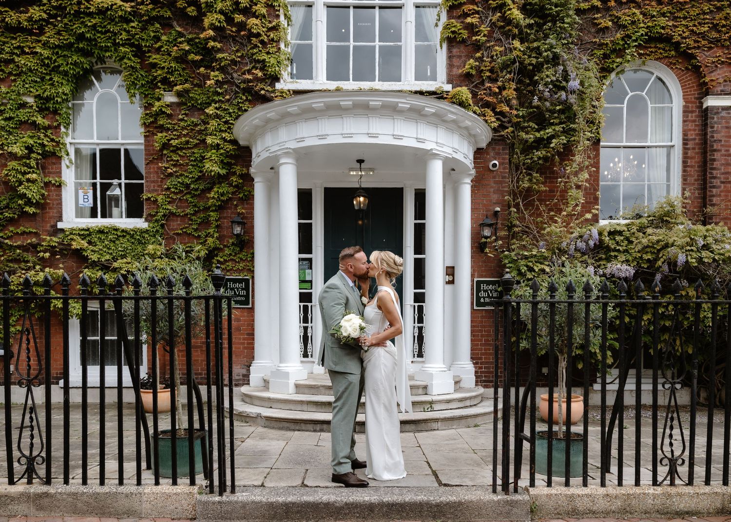 Bride and groom kissing outside ivy-covered brick wedding venue with white portico entrance and iron fence.