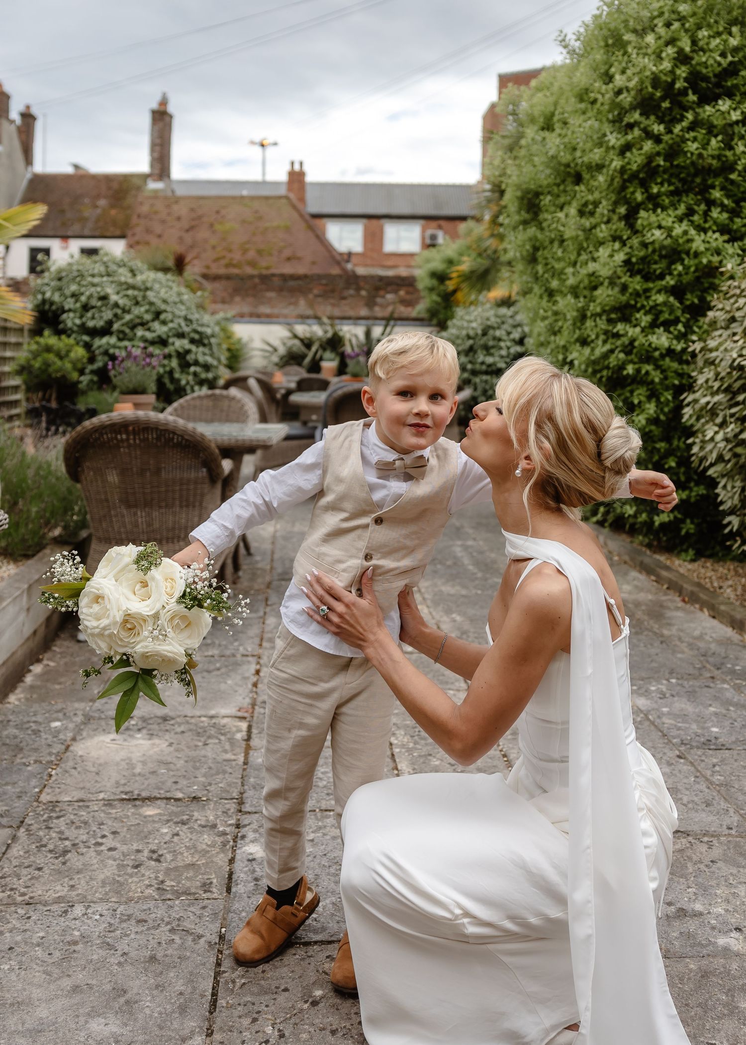 Blonde bride in white gown crouches to kiss young boy in beige suit holding white bouquet in garden.