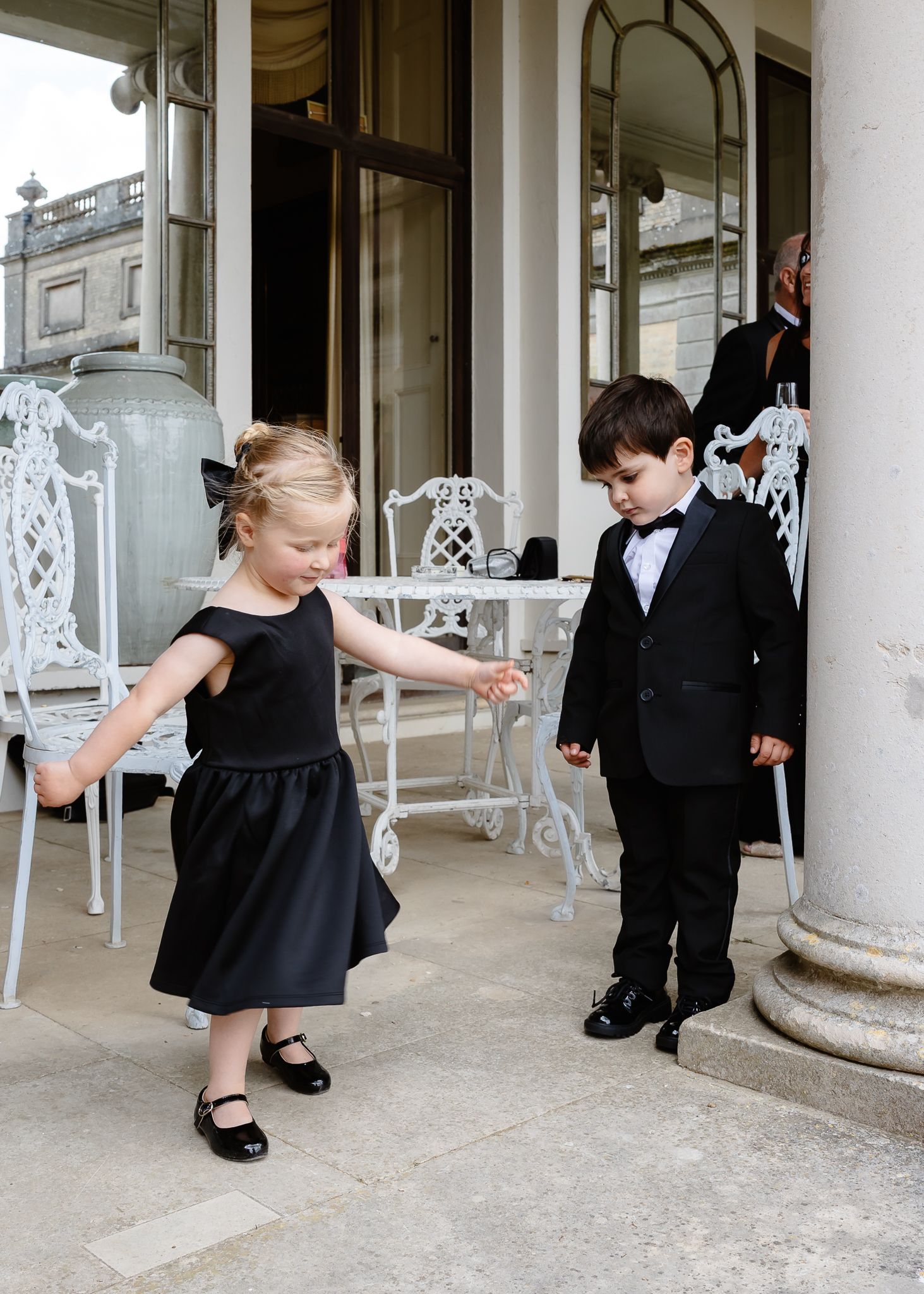 Two children in formal attire stand on an elegant porch with ornate white furniture and classical columns.